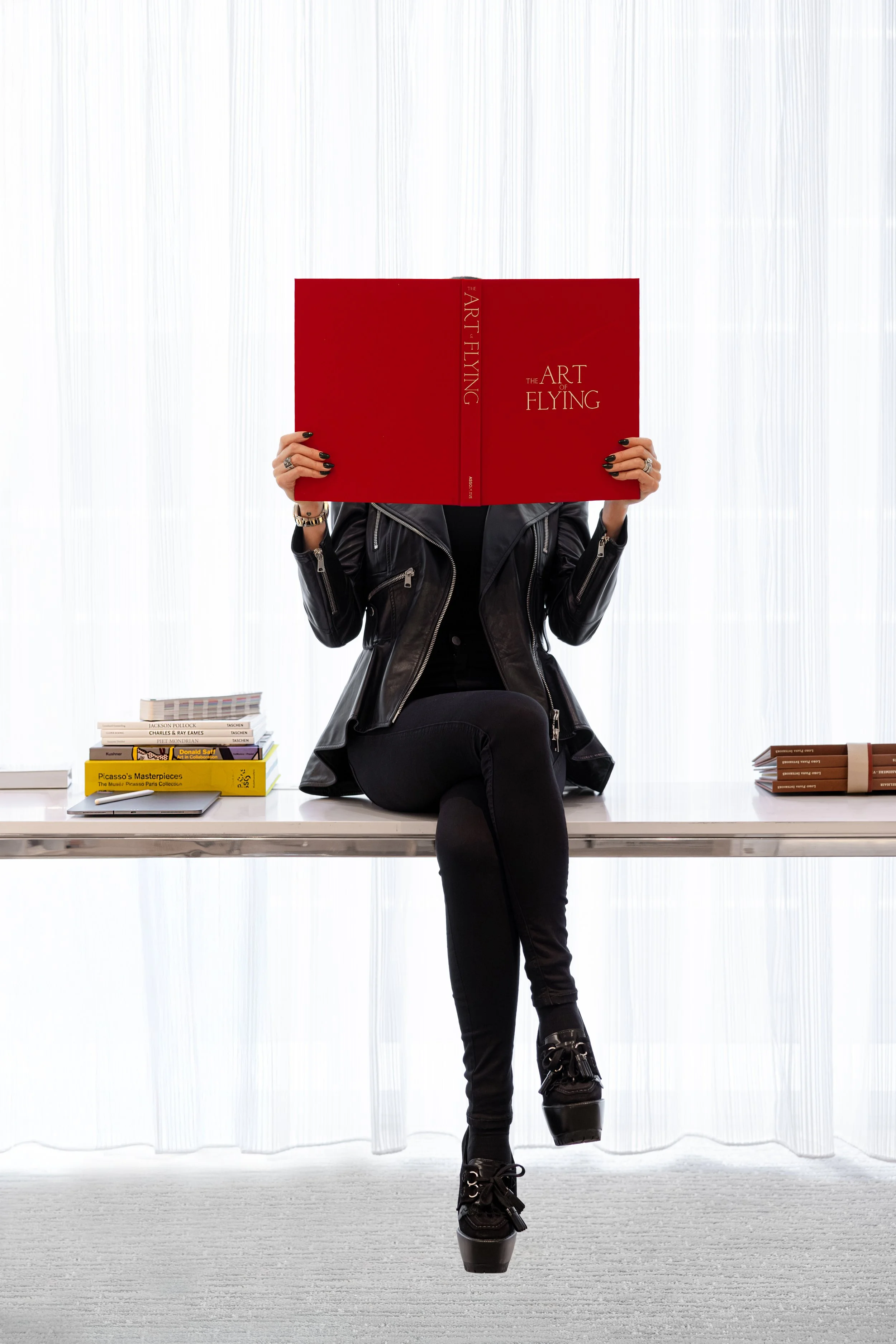 Top private jet designer Sarah Larranaga, holding a large red book titled "The Art of Flying," wearing a black leather jacket and black pants, with a stack of books on the table and a white curtain in the background.