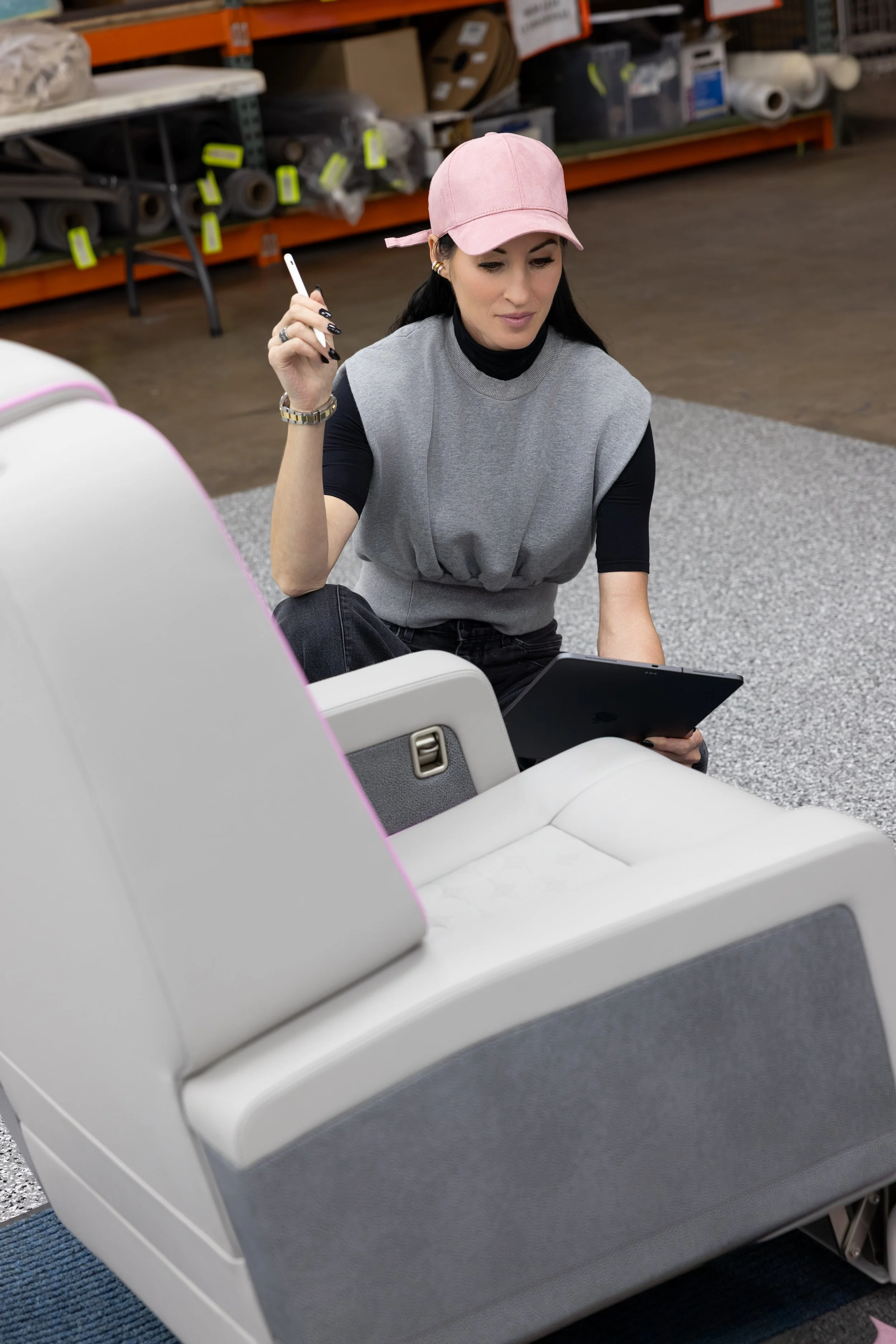 Top private jet designer Sarah Larranaga, wearing a pink cap, a gray sleeveless top over a black long-sleeve shirt, and gold jewelry, sitting on the floor of a hardware store with shelves of supplies behind her.