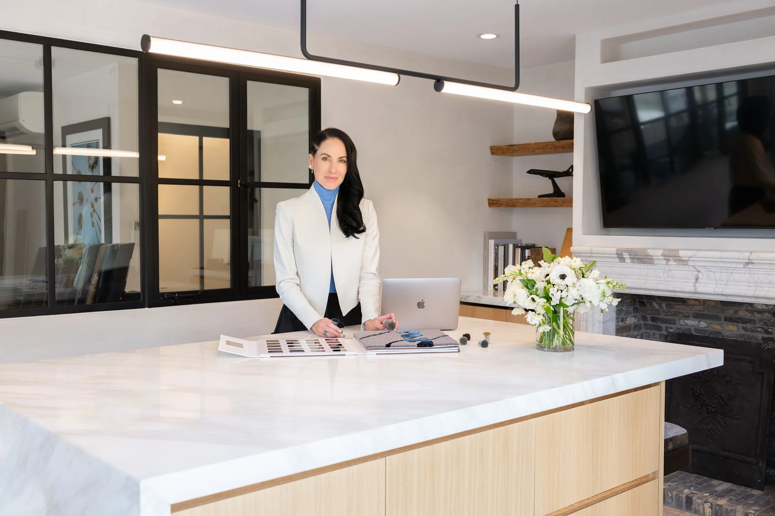 Top private jet designer Sarah Larranaga standing behind a white kitchen island with sample color swatches, fabric samples, and an open magazine, in a modern kitchen with glass partitions, a black TV, and a vase of white flowers.