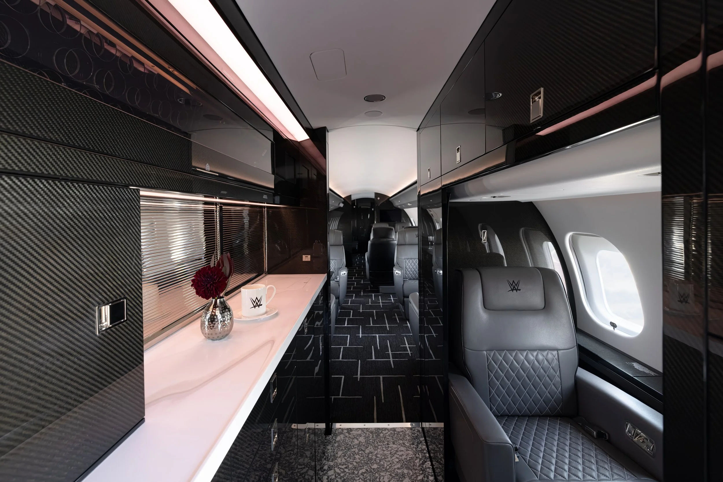Interior of a Bombardier Global 6500 by Sarah Larranaga Aviation Design with black and gray leather seating, a white countertop with a mug and a vase of flowers, and black glossy cabinets