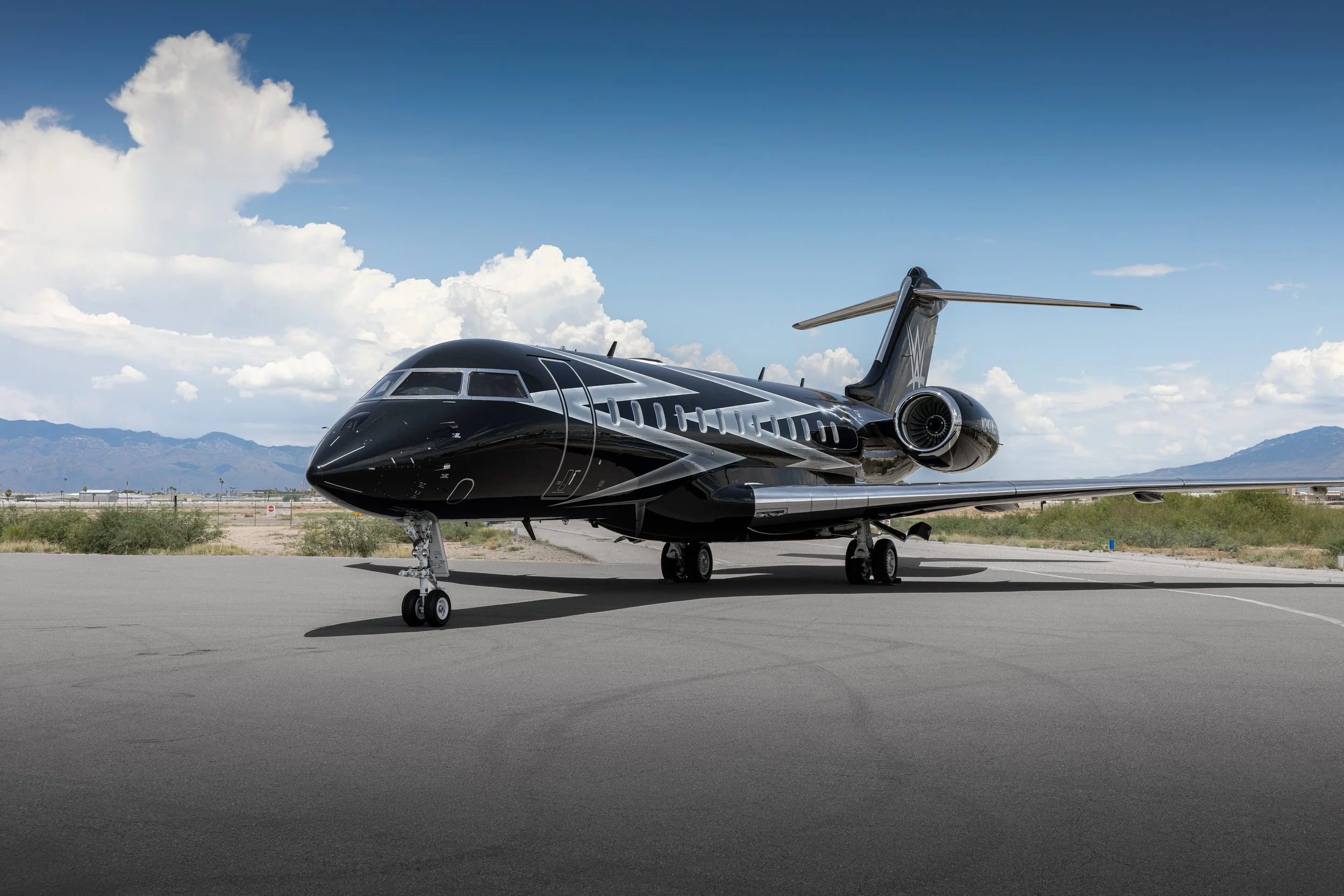A sleek black Bombardier Global 6500 by Sarah Larranaga Aviation Design private jet parked on an airstrip with a mountainous landscape and partly cloudy sky in the background.