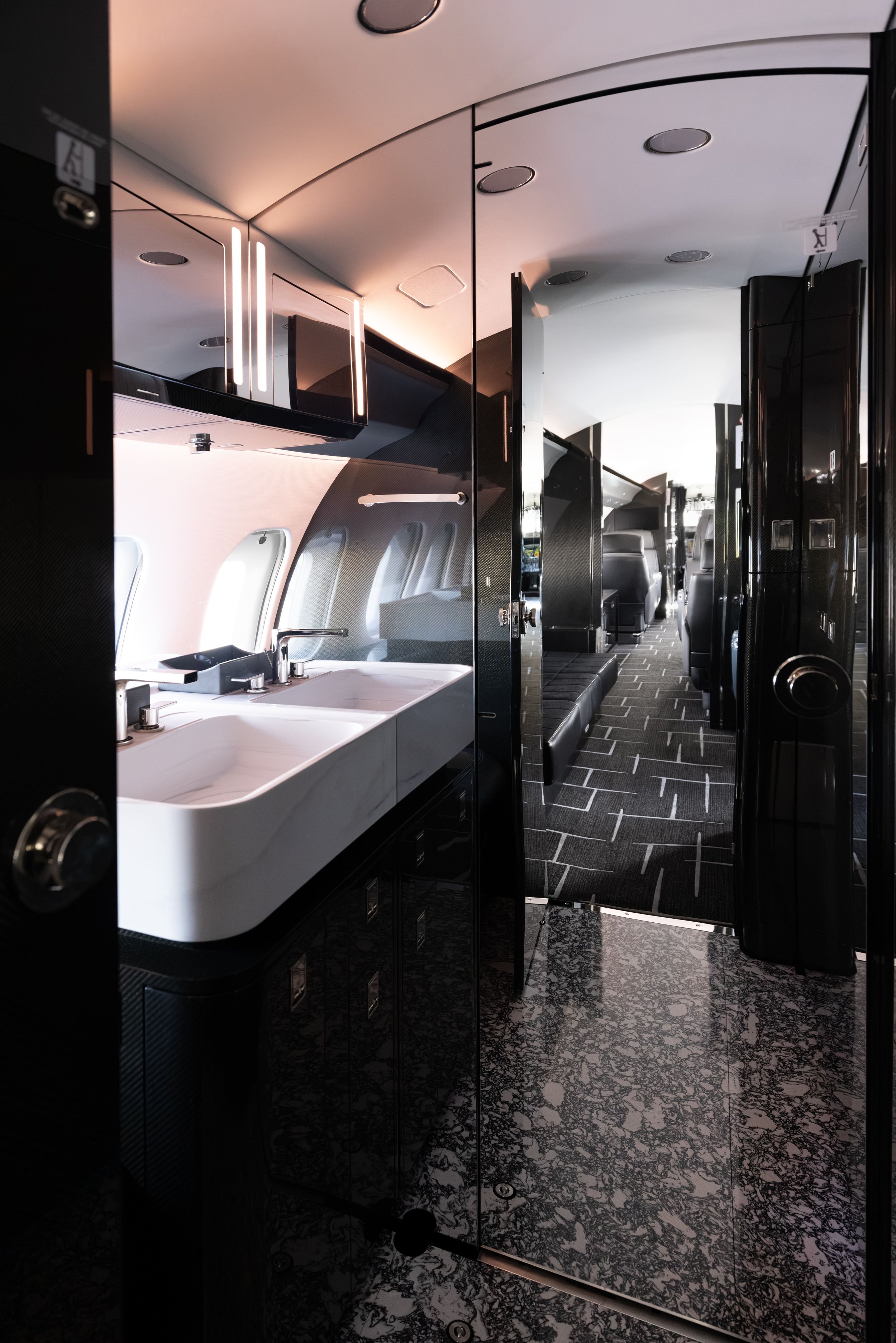 Interior of a Bombardier Global 6500 by Sarah Larranaga Aviation Design lavatory showing a black and white color scheme, a sink with a mirror above it, a partition door, and seating area visible in the background.