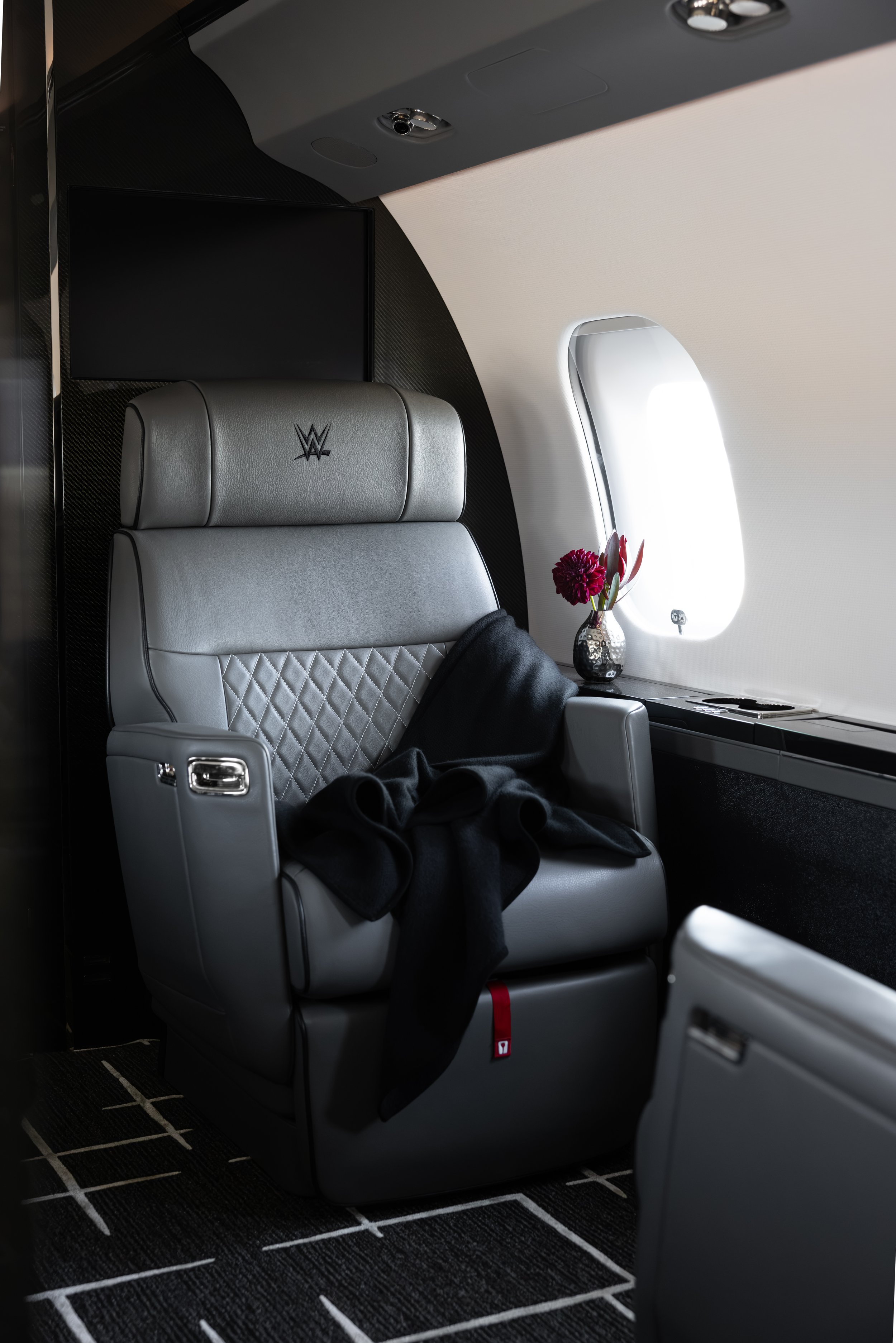 Inside a Bombardier Global 6500 by Sarah Larranaga Aviation Design with a gray leather seat featuring the WWE logo, a black blanket, a window with a flower vase, and overhead lighting.