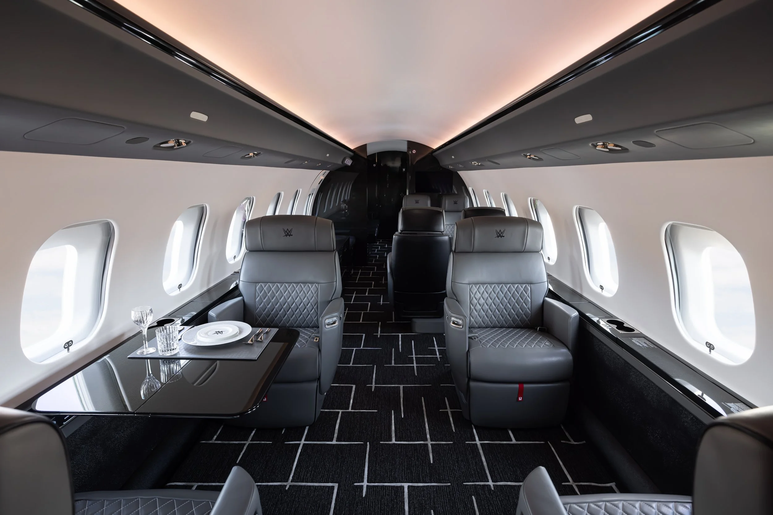 Inside of a Bombardier Global 6500 by Sarah Larranaga Aviation Design with gray leather seats, a dining table set with plates and glassware, and large windows, featuring modern black accents and a sleek ceiling design.