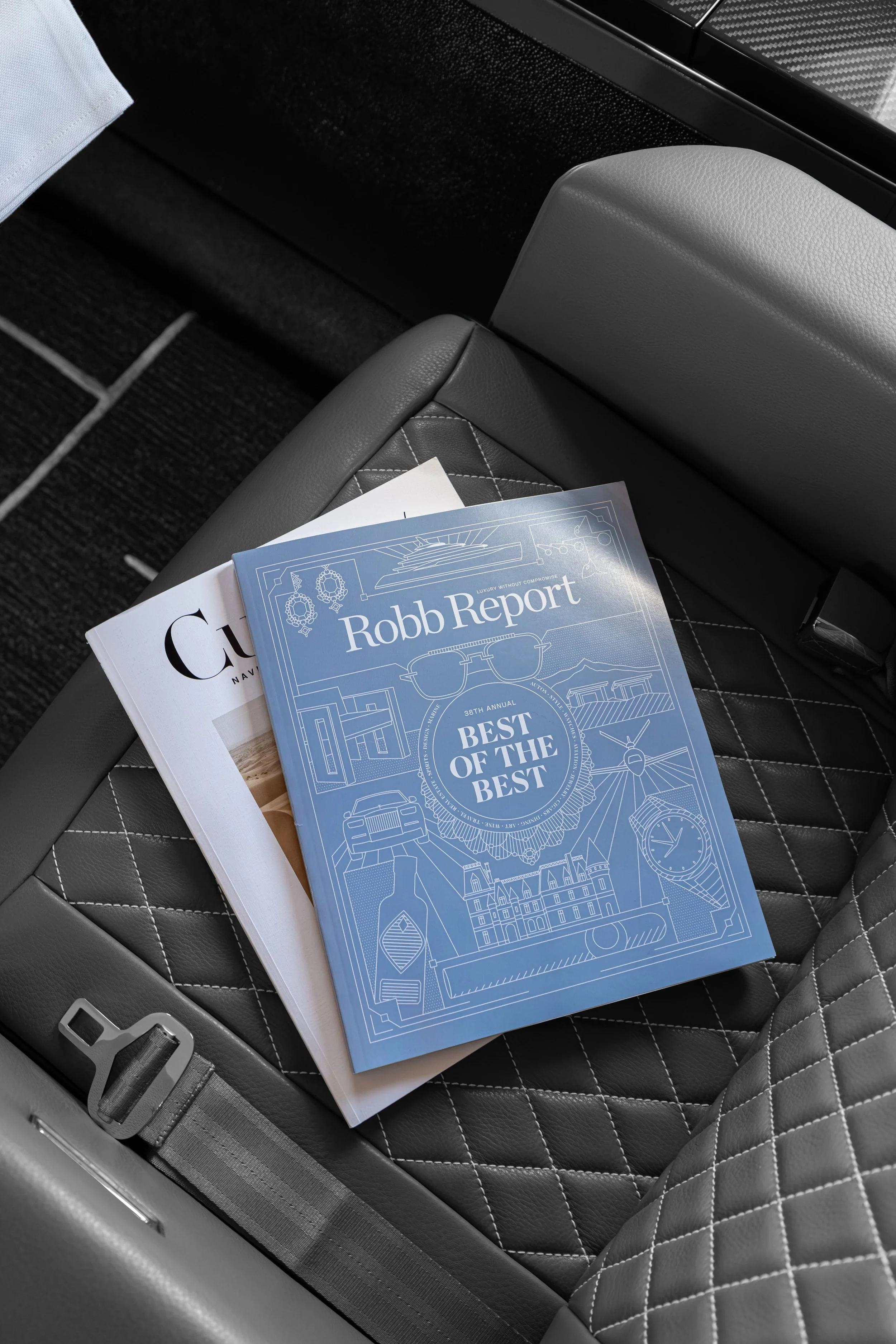An aircraft seat with a quilted pattern and gray color, holding pamphlets for Robb Report and The Curator publication.