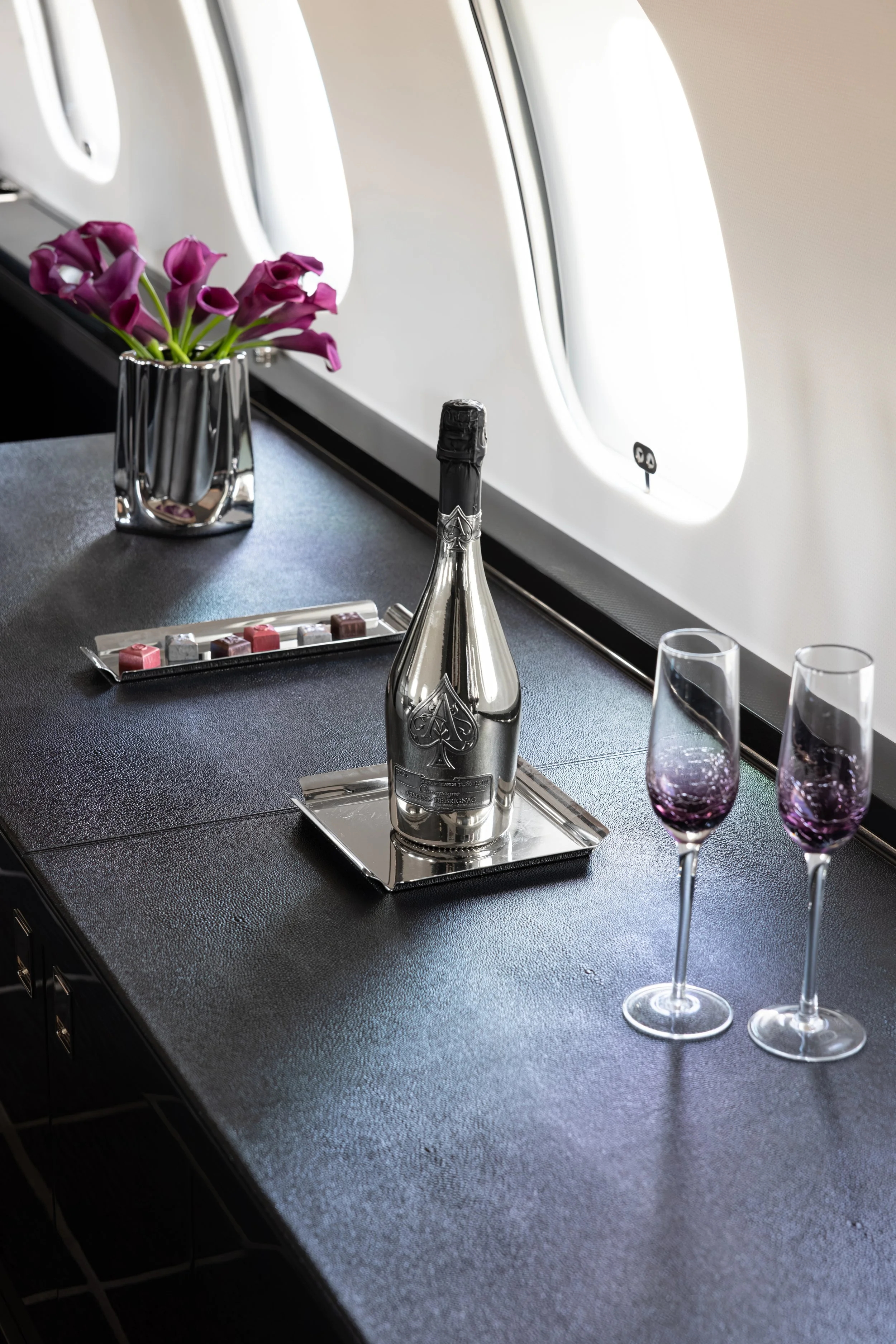 A table with a silver champagne bottle on a tray, two purple wine glasses, a small tray of chocolates, and a silver vase with purple calla lilies, inside an airplane cabin near a window.