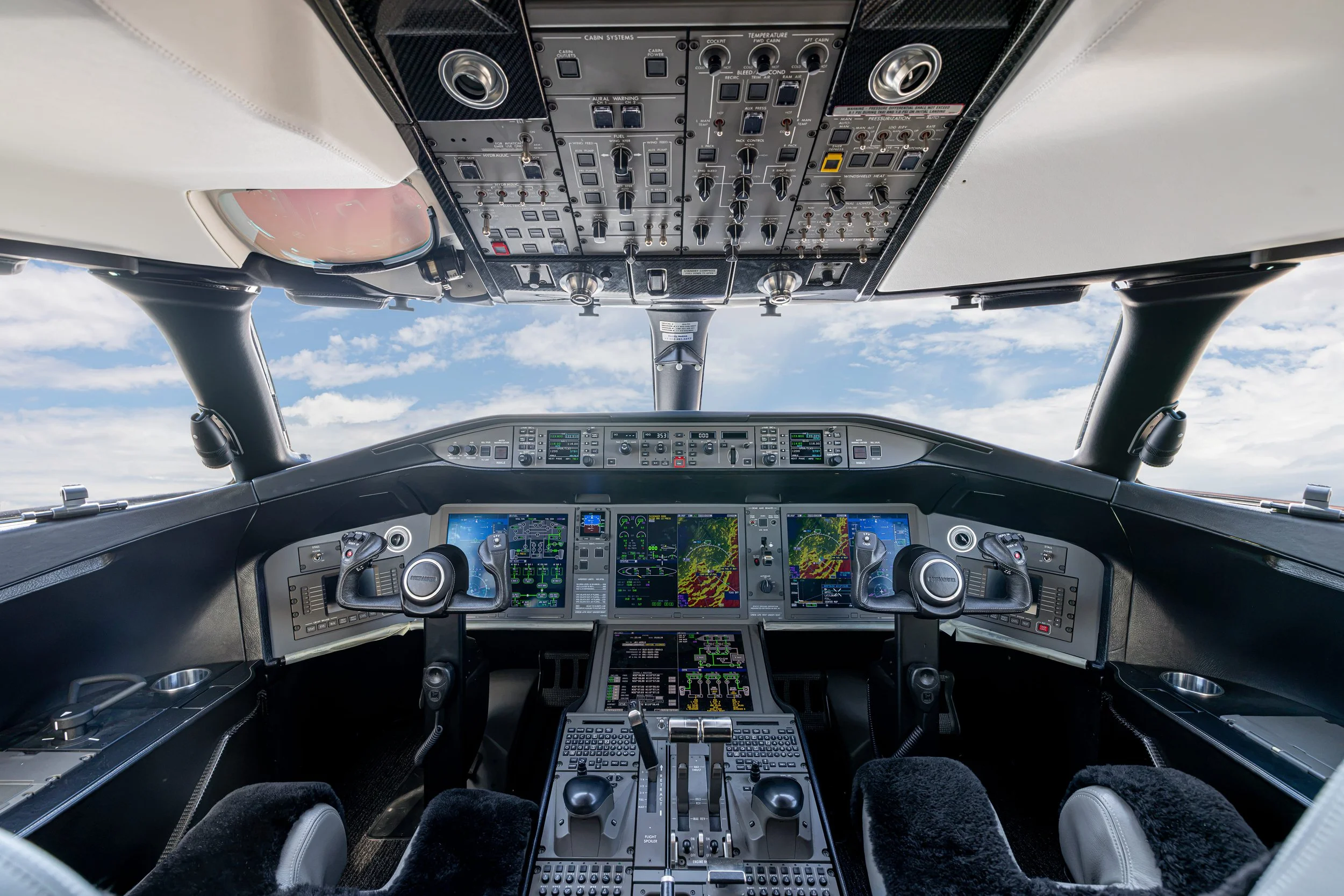 Bombardier Global 6500 by Sarah Larranaga Aviation Design. Pilot cockpit with multiple screens, control panels, and instrumentation, with clouds visible through the windshield.