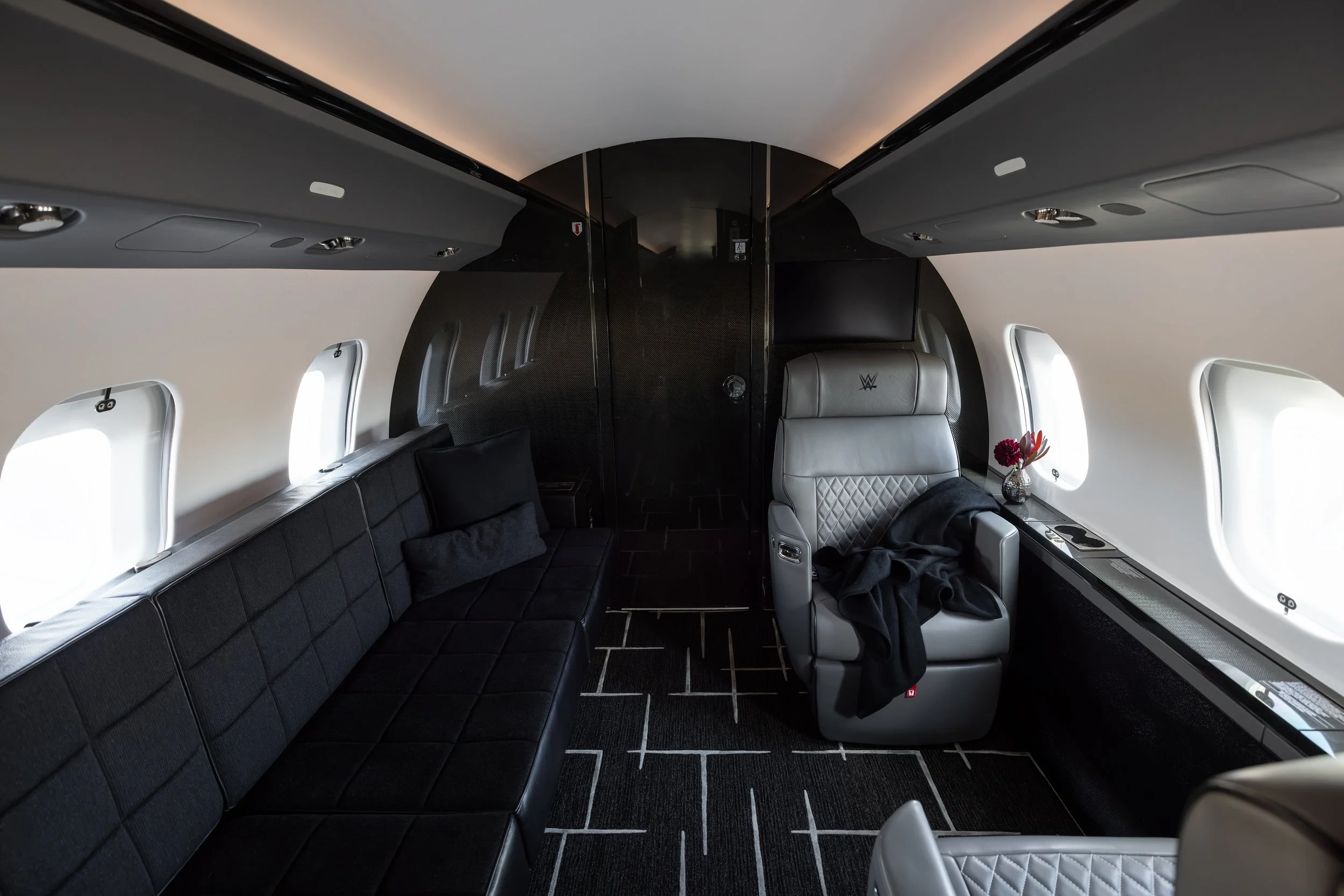 Inside a Bombardier Global 6500 by Sarah Larranaga Aviation Design with black leather seating, glass windows, a small table with flowers, and a television screen.
