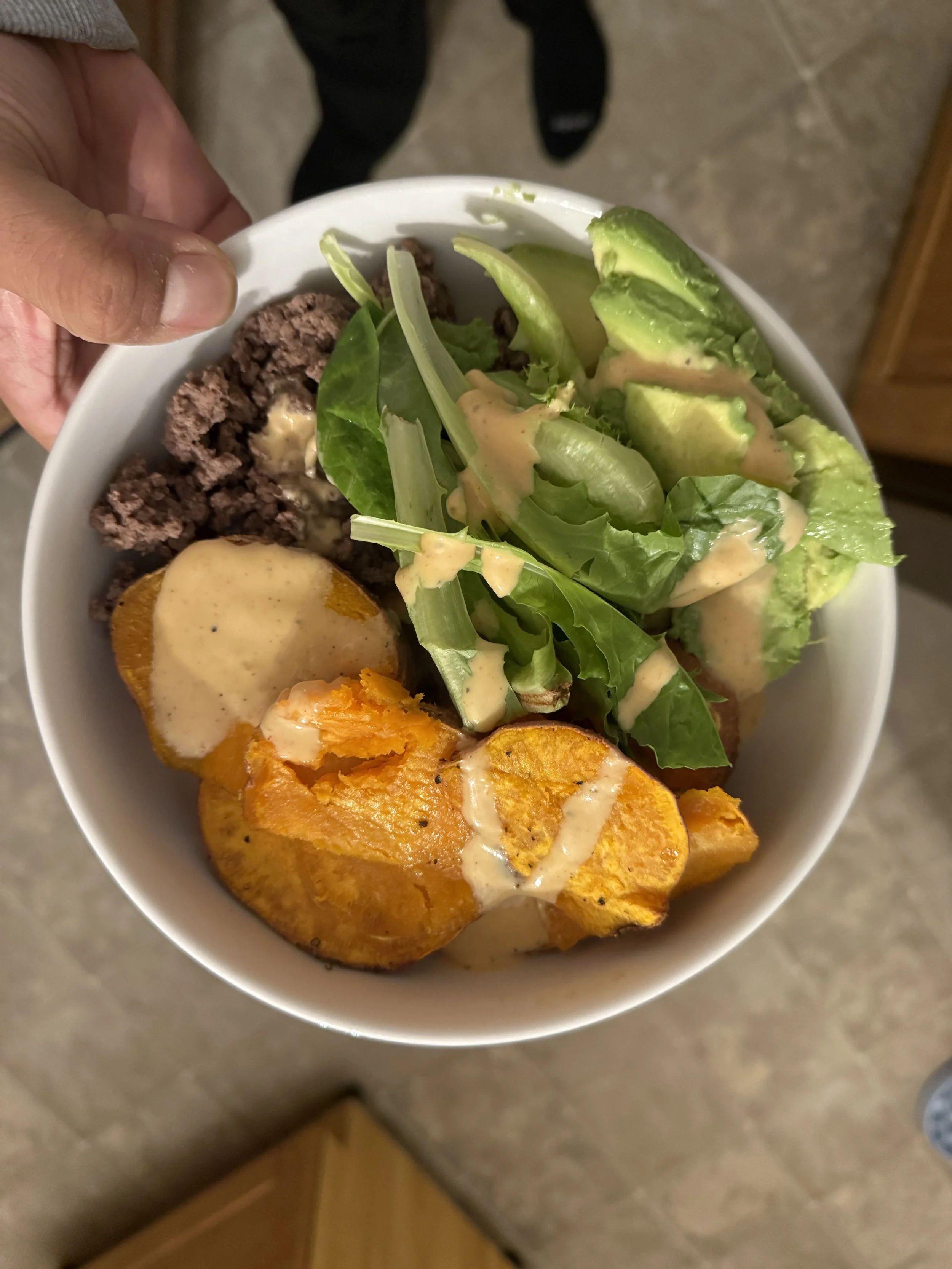 A bowl containing a salad with fresh greens and sliced avocado, topped with a creamy dressing, pieces of roasted sweet potato with dressing, ground beef, and black beans.