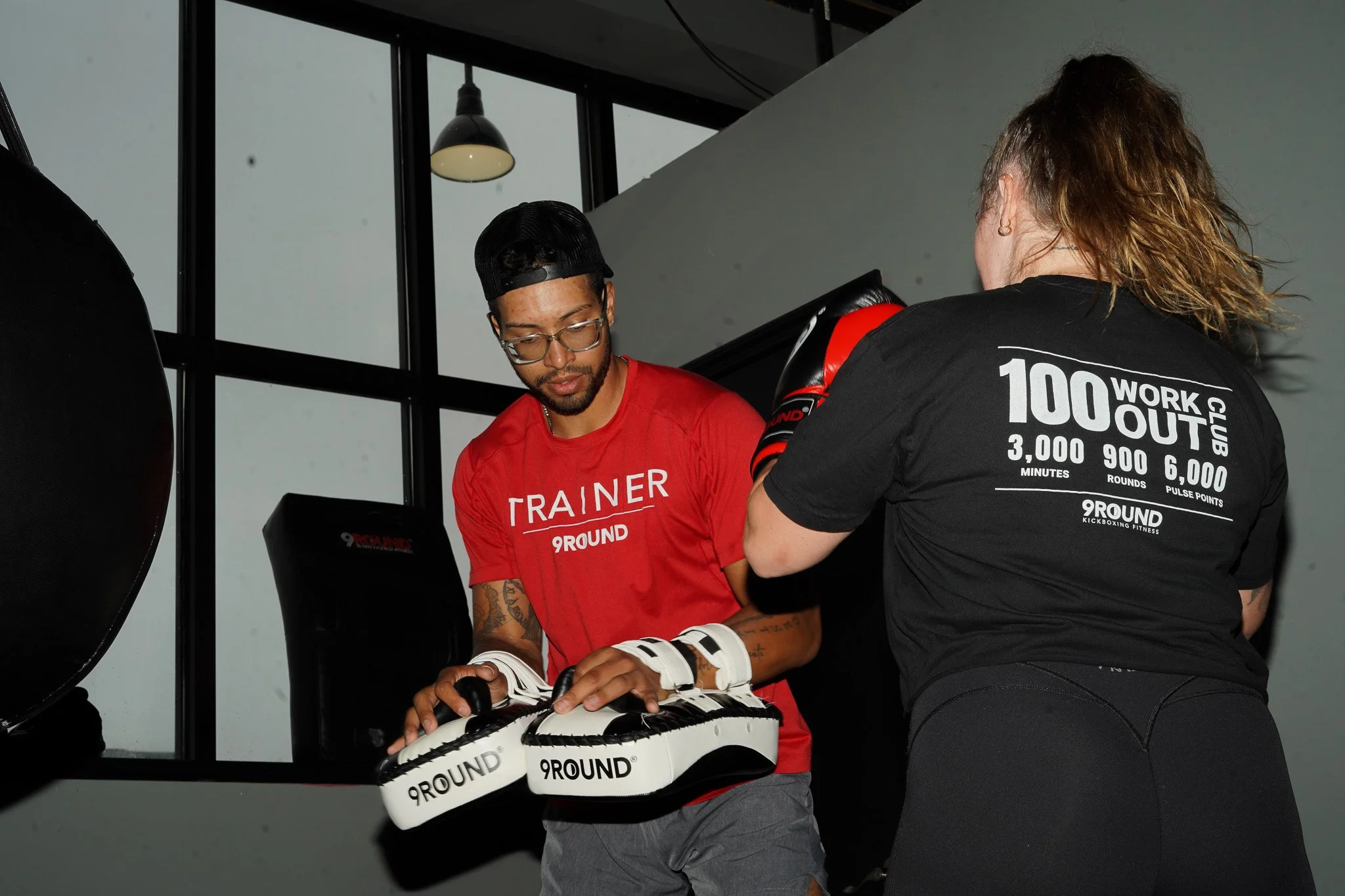 A fitness trainer and a woman with curly hair working out at a gym. The trainer is holding padded gloves branded '9ROUND'. The woman is preparing to punch with boxing gloves, and she is wearing a black shirt with workout information on the back.