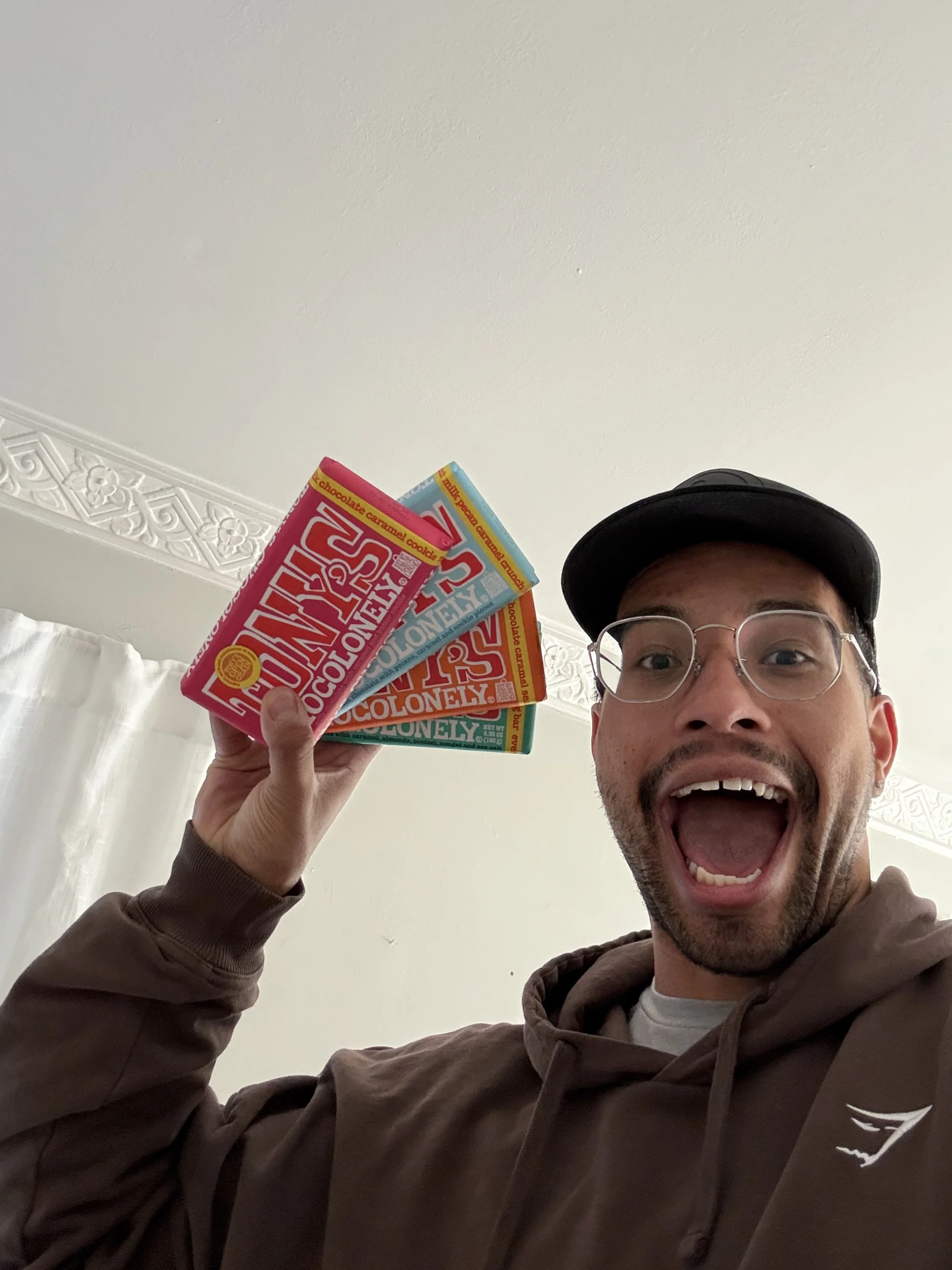 A man wearing glasses, a cap, and a brown hoodie holding three colorful chocolate bar wrappers, smiling with an open mouth.