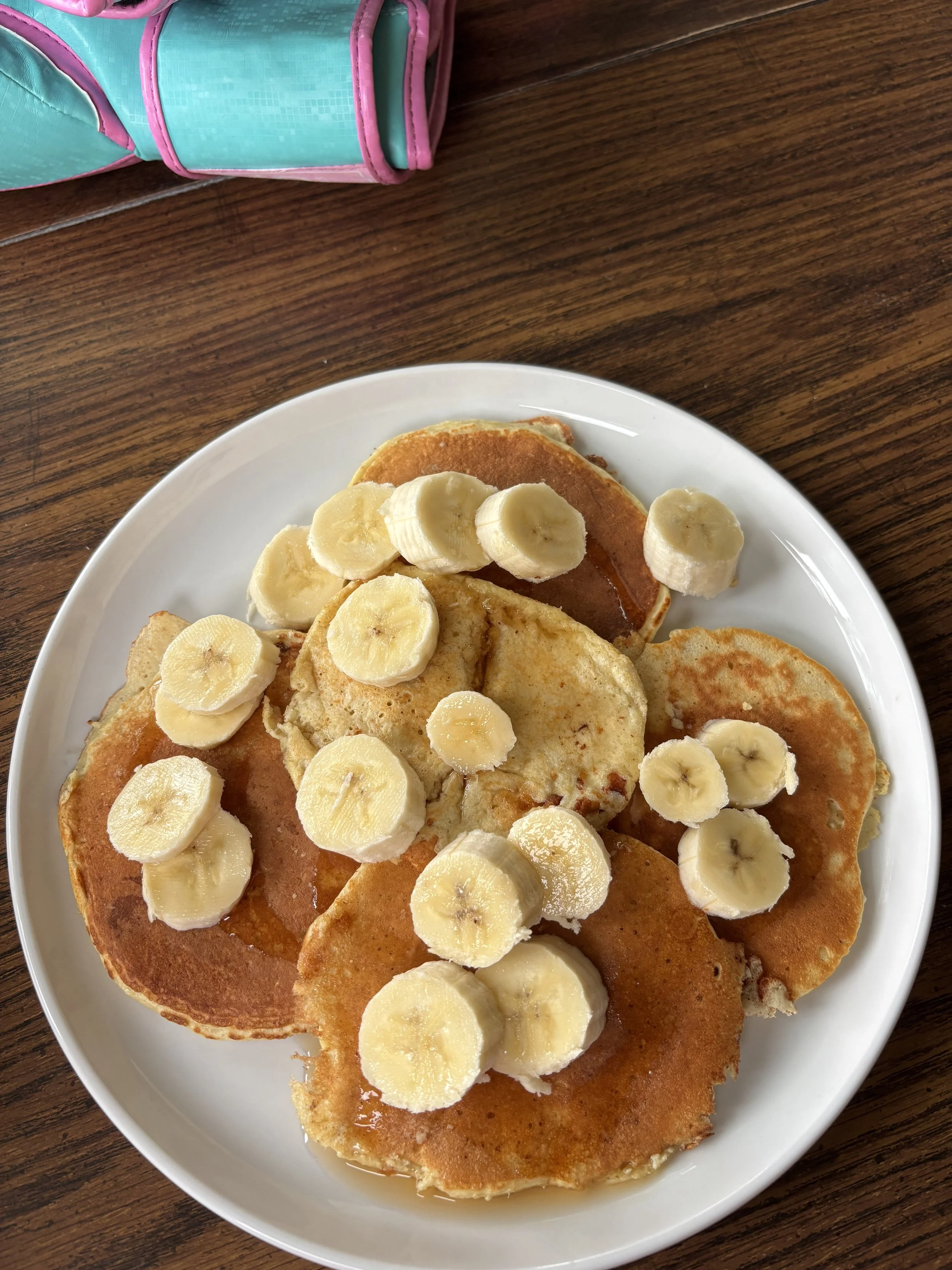 Plate of banana and pancake slices on a white dish, on a wooden table with a pink and teal bag in the corner.