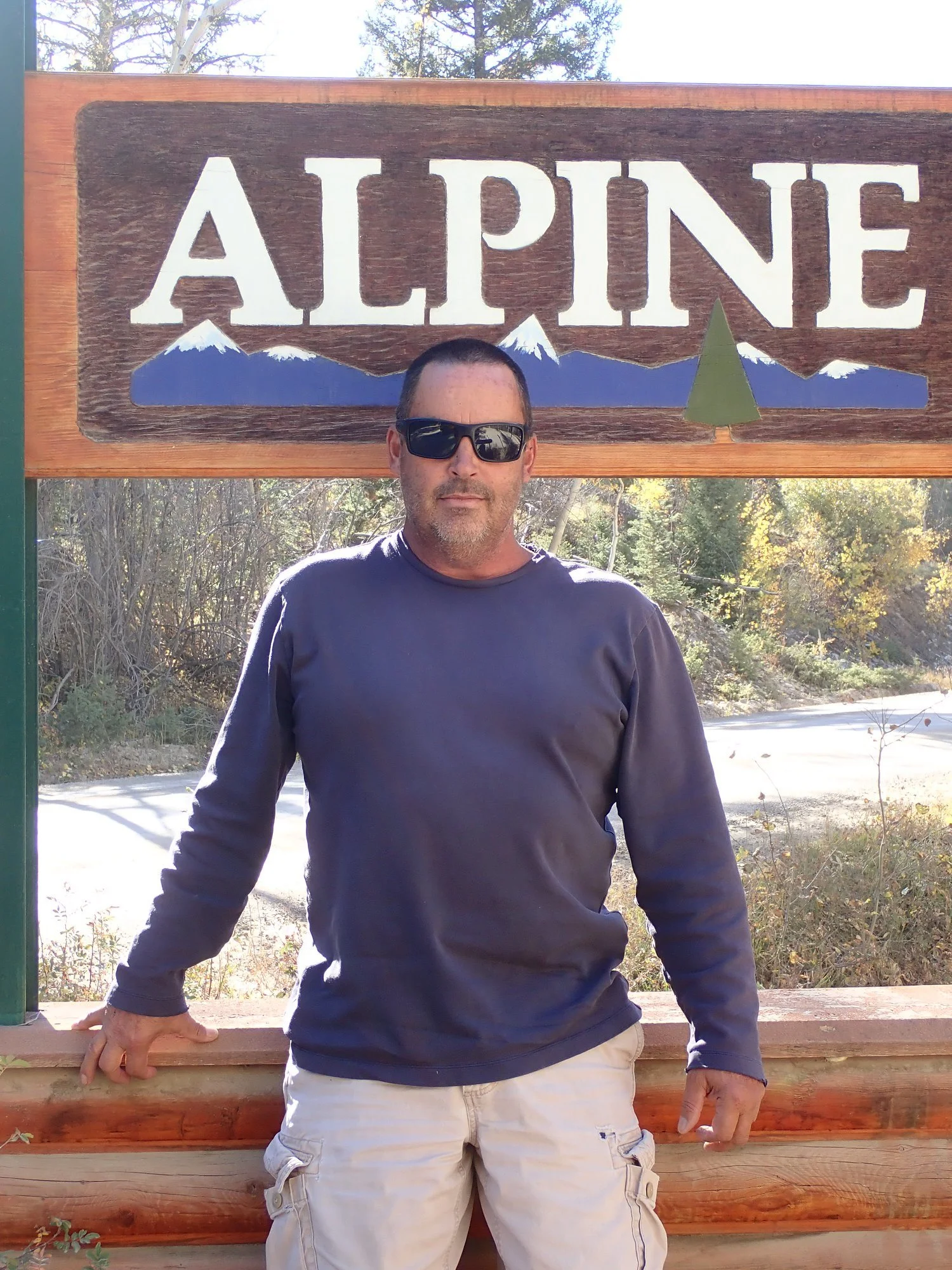 Person wearing sunglasses and a dark long-sleeve shirt standing in front of a sign that says 'Alpine' with mountains and a tree in the background.