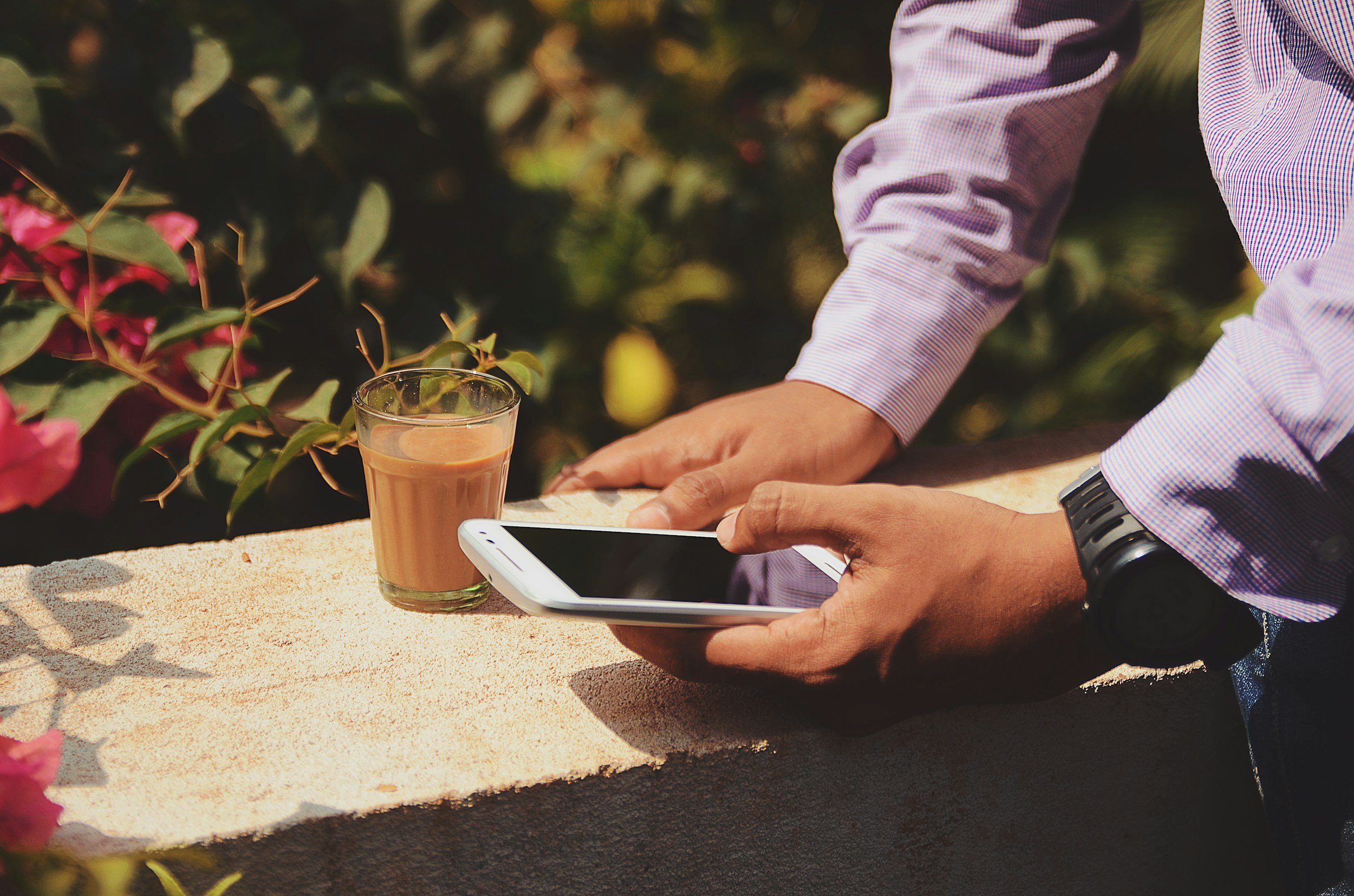 Person holding a smartphone next to a glass of tea on a ledge, with foliage in the background.