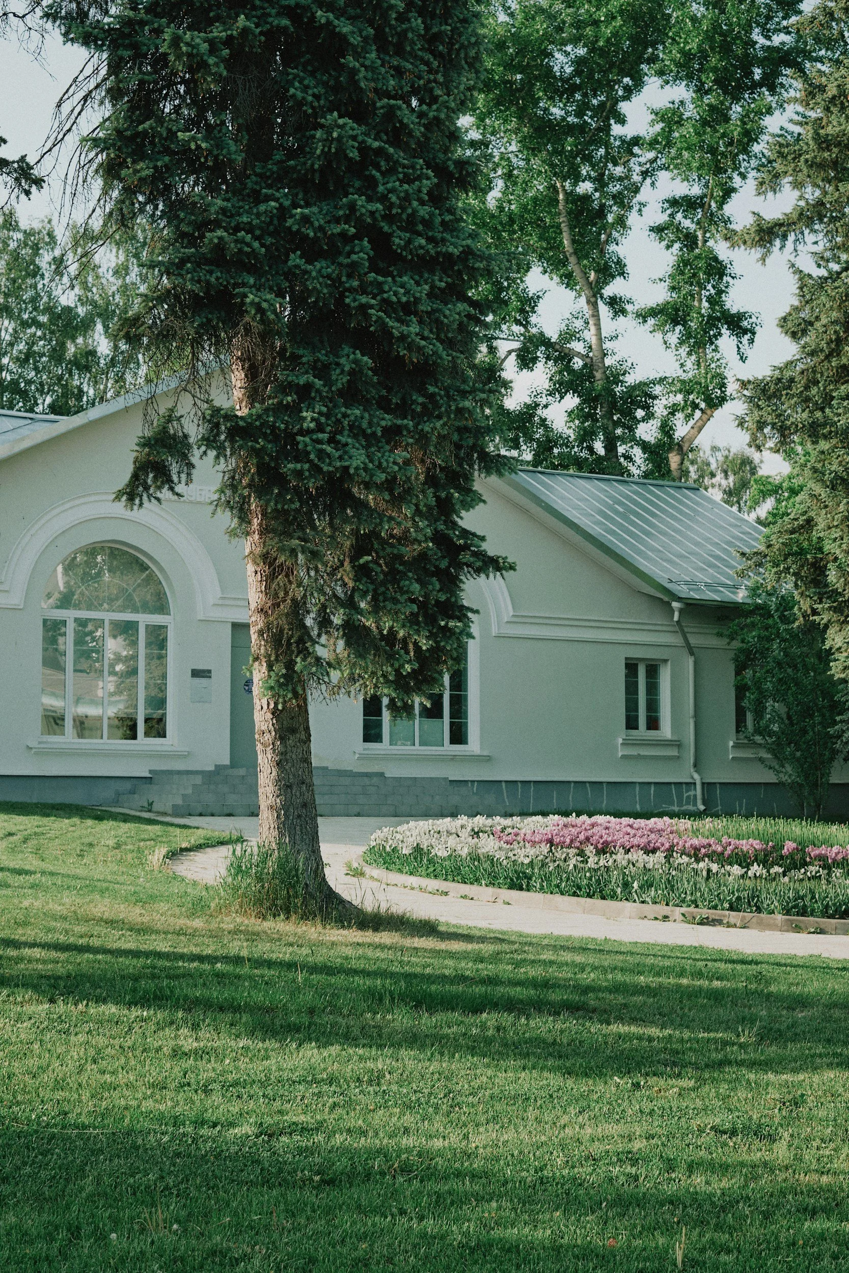A white building with arched windows surrounded by trees and a garden with colorful flowers, set against a green lawn.