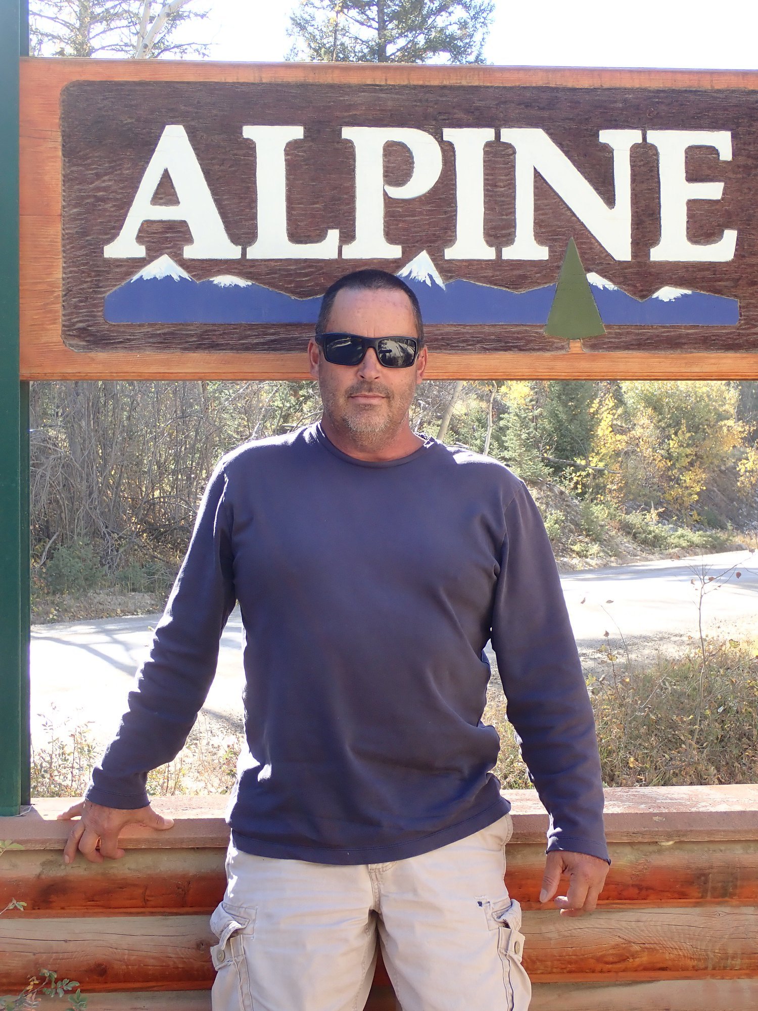 A man wearing sunglasses standing in front of a wooden sign that says "ALPINE" with a mountain silhouette and tree illustration.