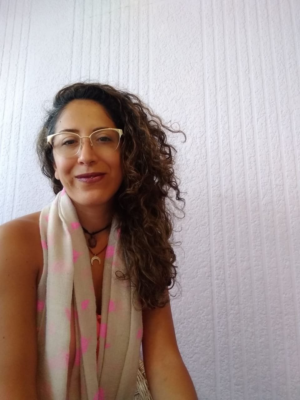 Mujer con gafas, bufanda y collar, sonriendo en una pared de textura blanca.