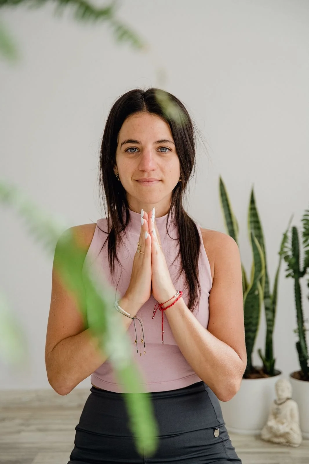 Mujer joven con manos juntas en actitud de oración o gratitud, de pie en un espacio con plantas de interior y fondo claro.