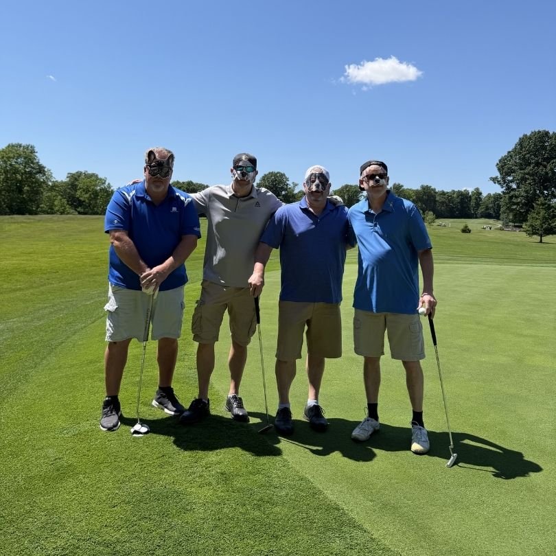 Four men on a golf course wearing sunglasses and camouflage face paint, holding golf clubs, standing on the green under a blue sky with a couple of clouds.