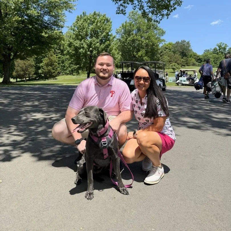 A man and a woman are squatting next to a black Labrador wearing a pink harness on a golf course with golf carts and people in the background. They are outdoors on a sunny day, surrounded by green trees.