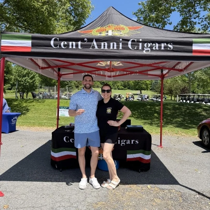 Two people standing under a black canopy with 'Cent' Anni Cigars' written on it, smiling at the camera on a sunny day in a park with trees and golf carts in the background.