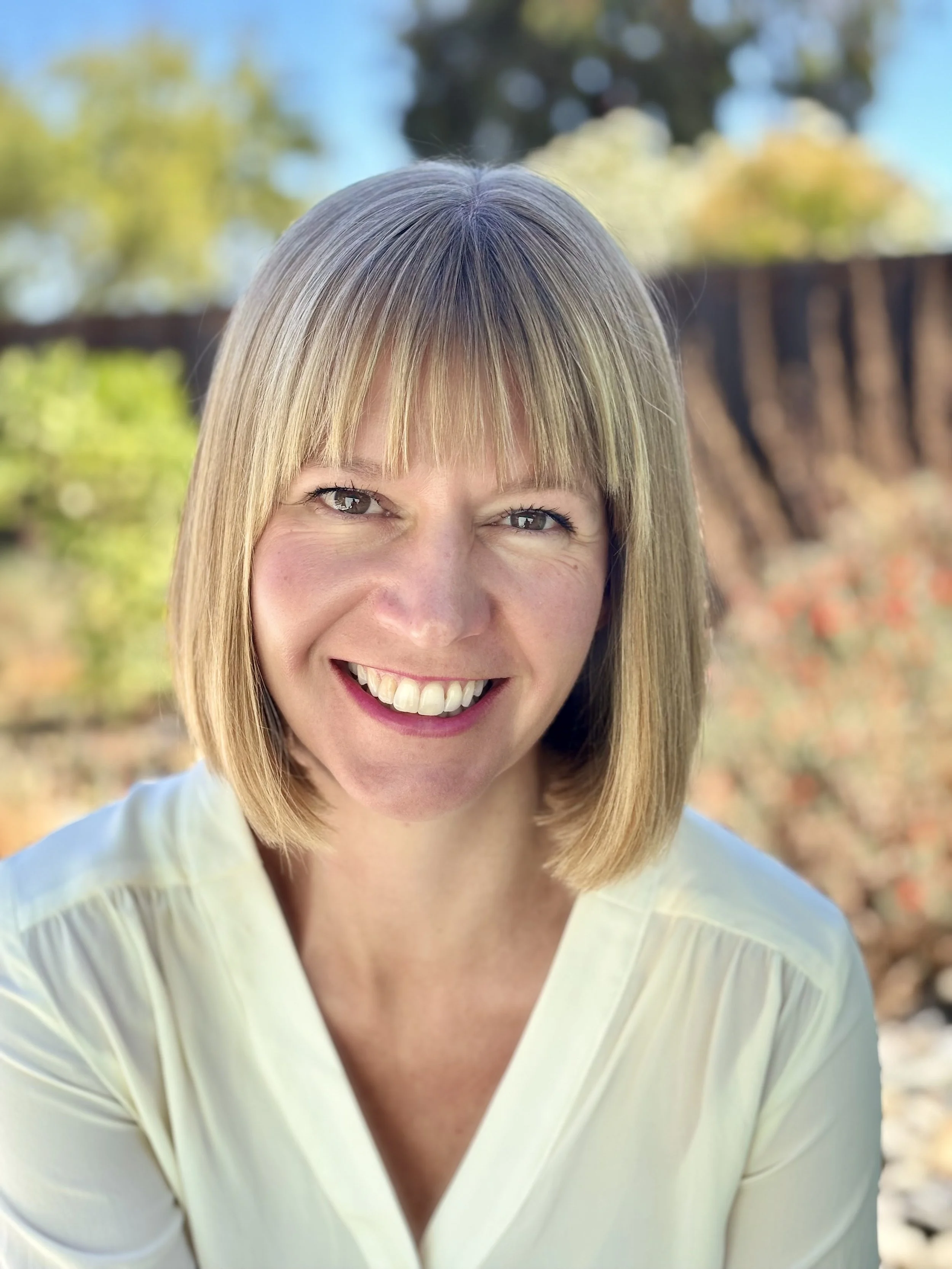 A woman smiling outdoors with trees and blurred background, wearing a light yellow shirt.