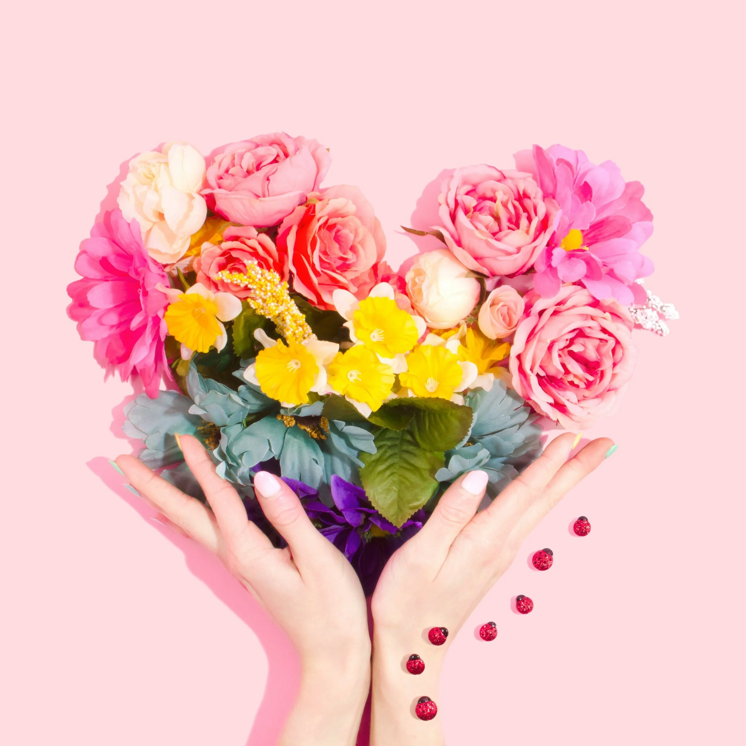 A pair of hands holding a colorful bouquet of pink, yellow, and white flowers with small red berries on a pink background.