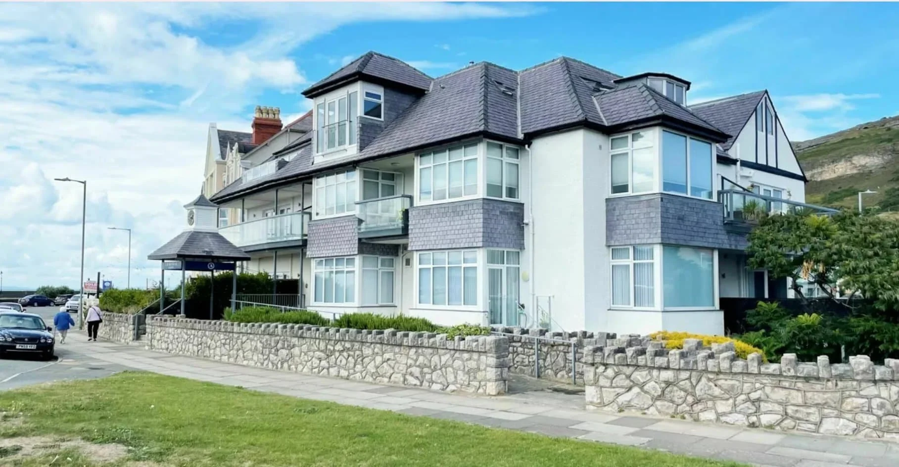 Modern, multi-storey white building with grey roof, stone wall, and sidewalk on a sunny day with blue sky.