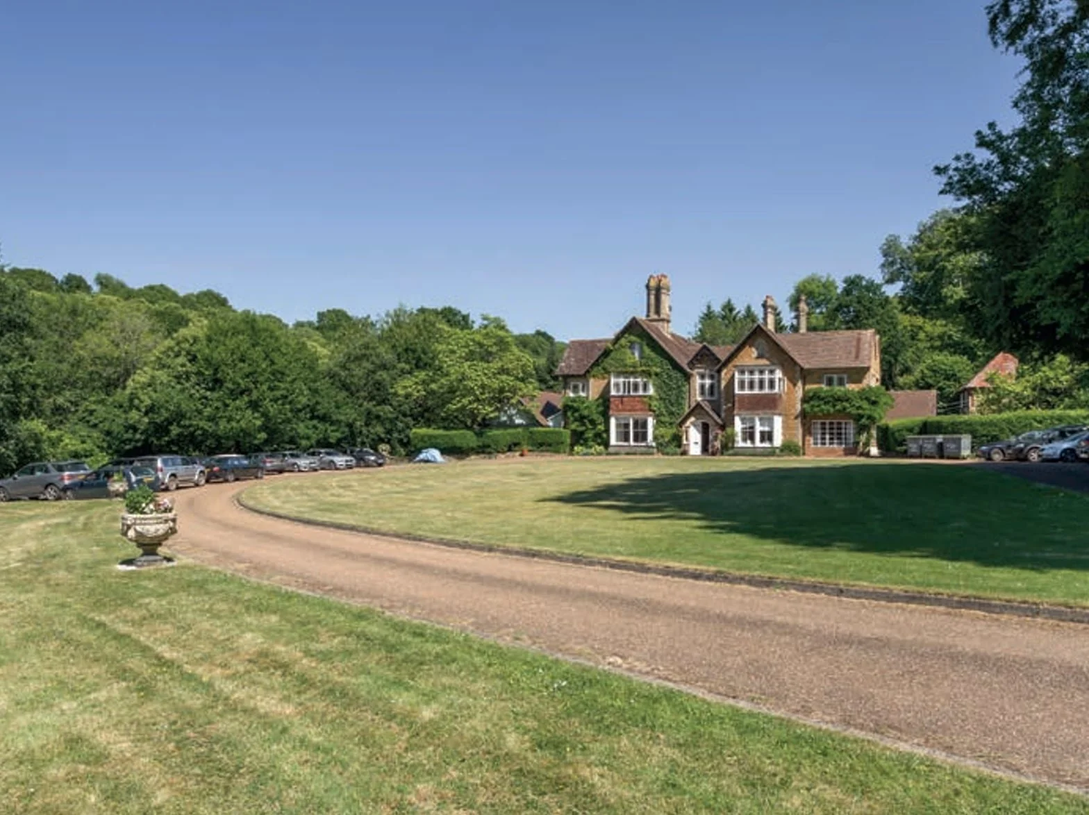 A large country house with a circular driveway, surrounded by trees and parked cars on a sunny day.
