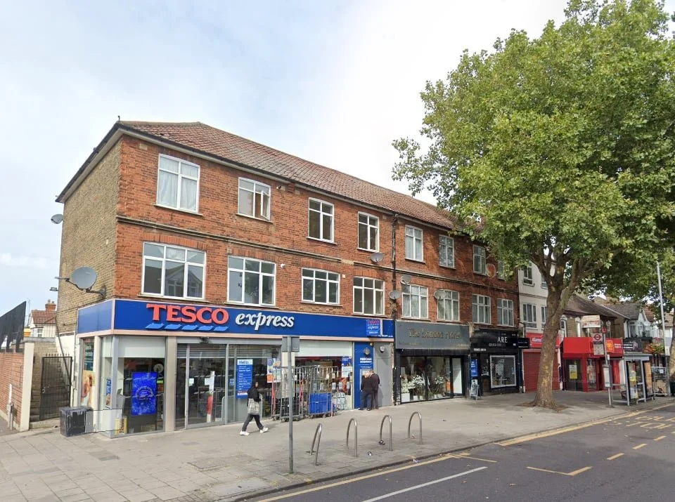 A Tesco Express store with shops and apartments above, located on a street with a tree and bicycle racks on the sidewalk.