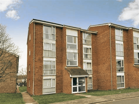 A three-story red brick apartment building with large windows and an entrance porch, set on a grassy lawn under a blue sky.