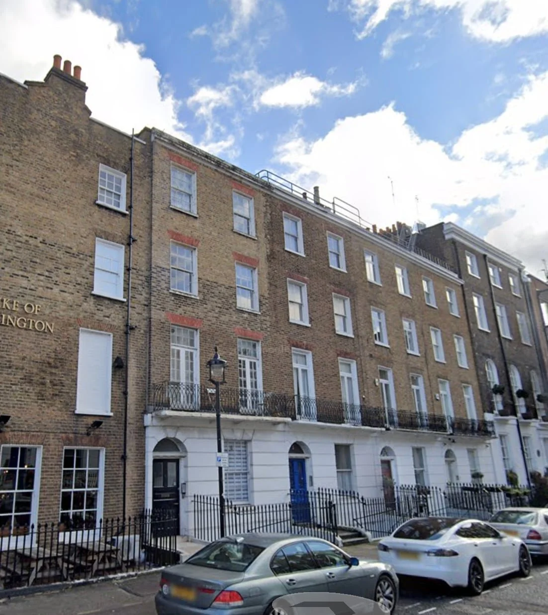 A row of Georgian-style terraced houses with brick facades and white trim, parked cars along the street, under a partly cloudy sky.
