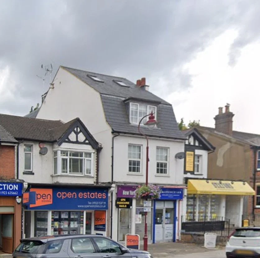 A row of shops with apartments above, including open estates, on a cloudy day with parked cars in front.