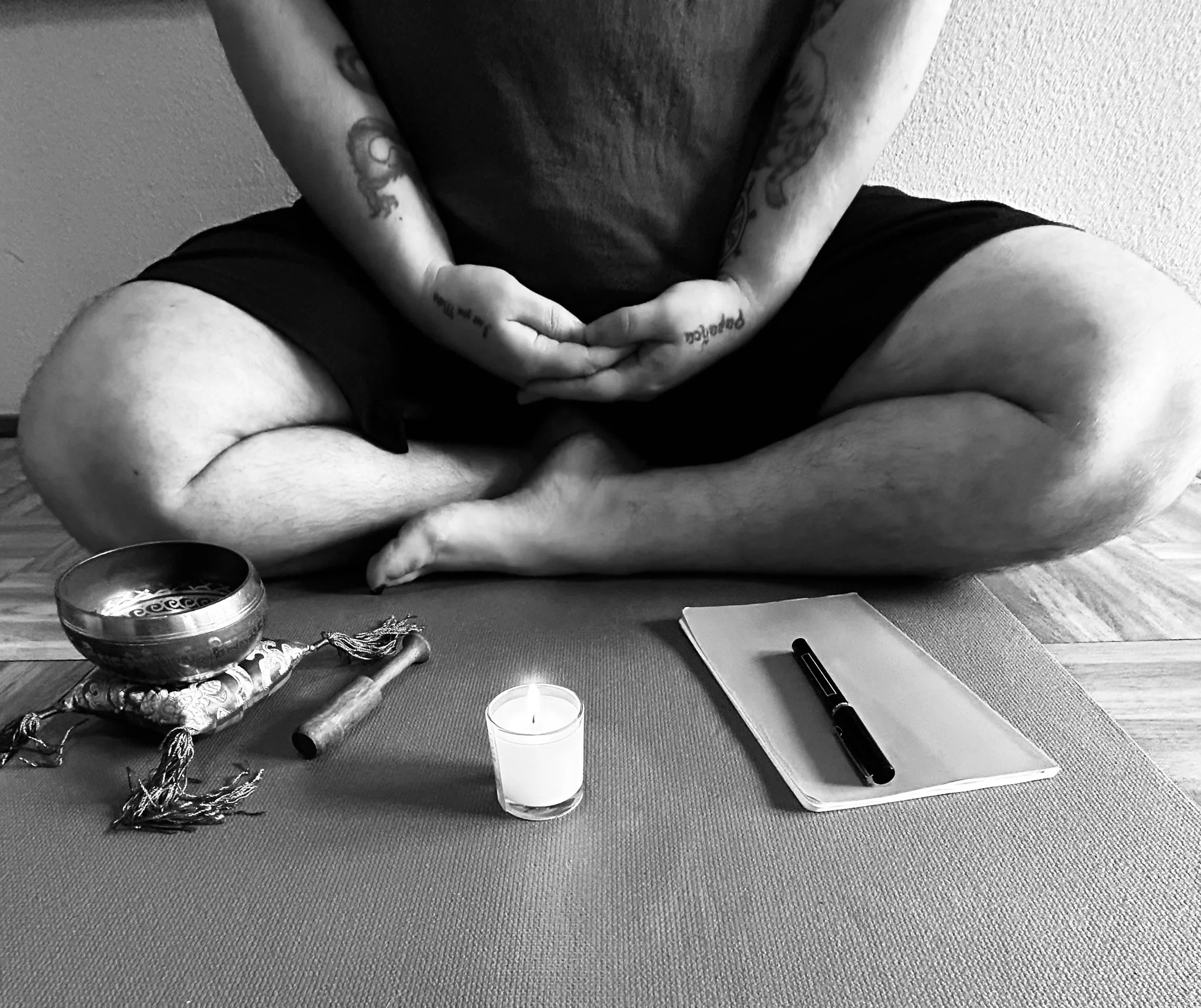 A person sitting cross-legged on a mat, with a candle, a journal with a pen, and a singing bowl in front of them. The person has tattoos on their arms and torso and is wearing black shorts.