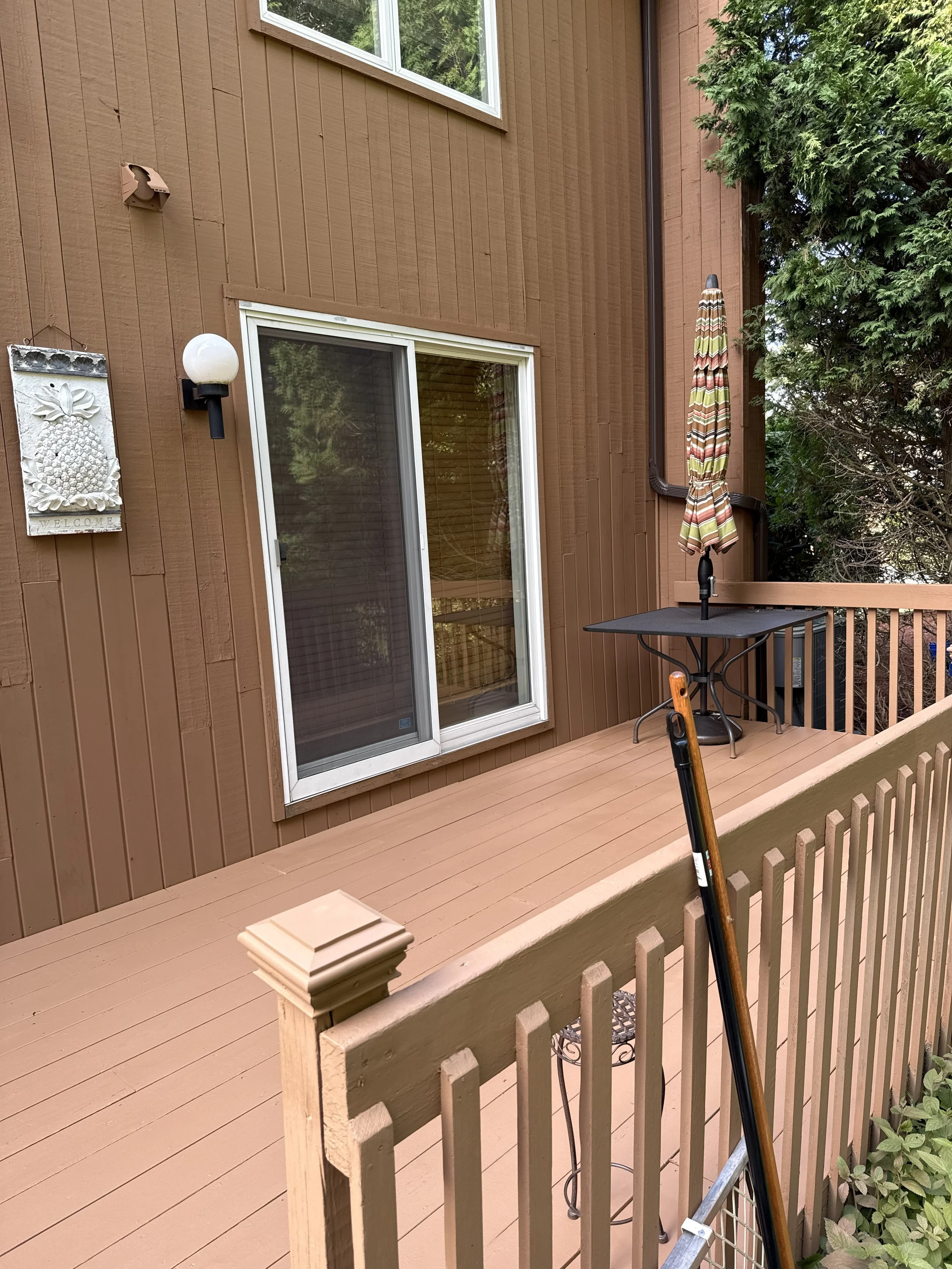 Wooden deck with patio furniture, umbrella, sliding glass door, outdoor lighting, and plants along the deck railing.