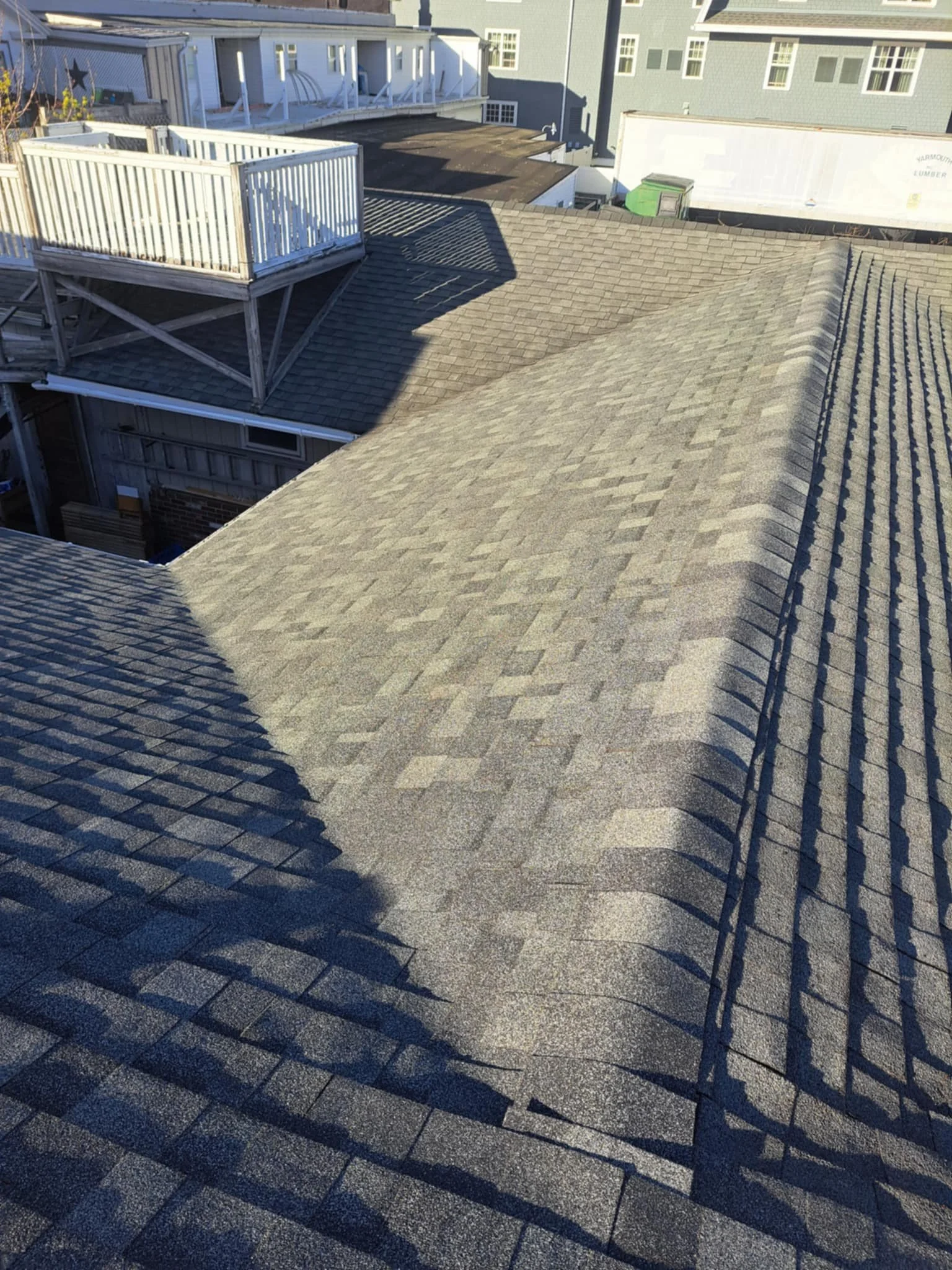A view of residential rooftops with asphalt shingles, showing shadows cast by a balcony and neighboring buildings.