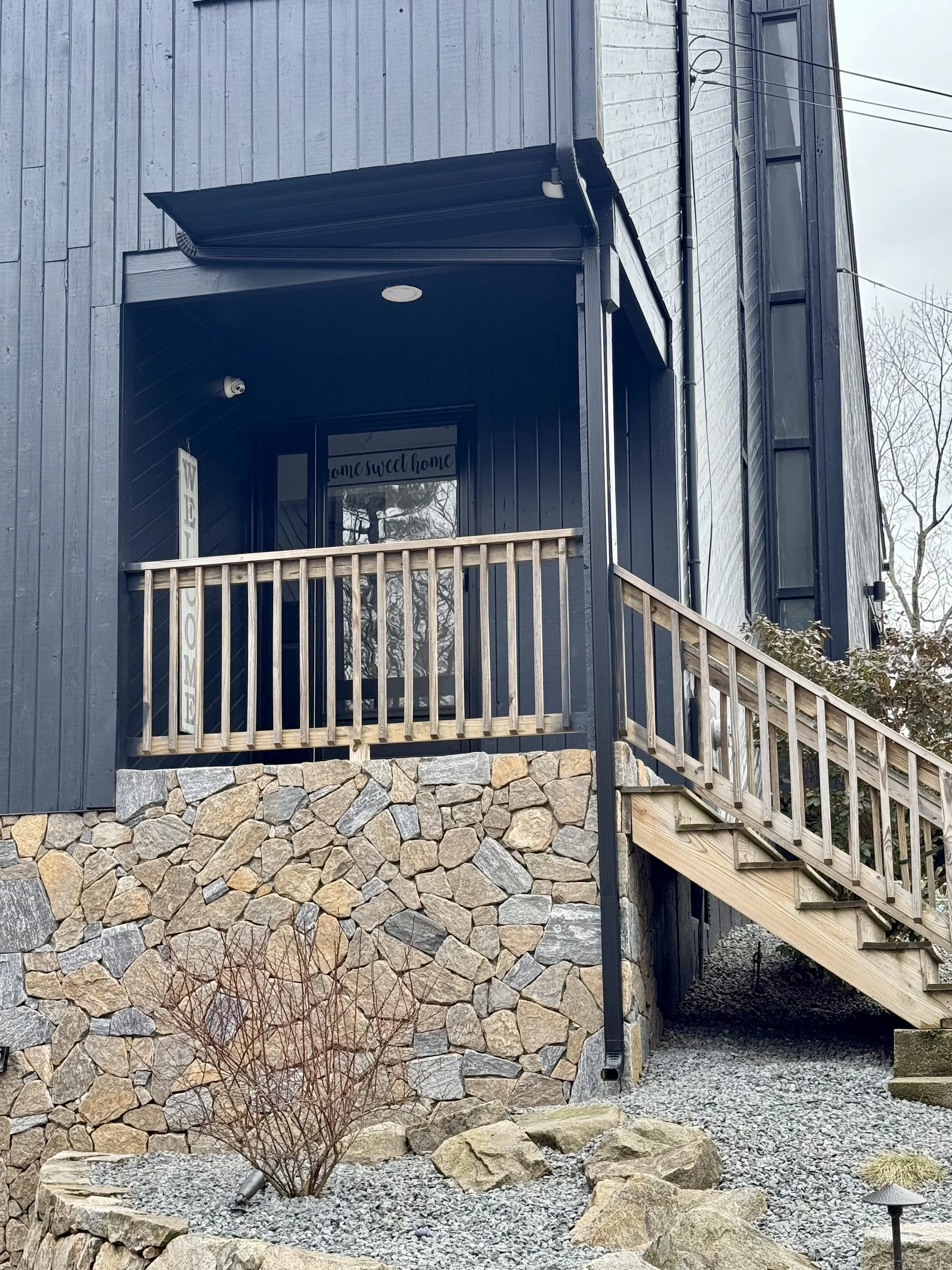 Front porch of a house with blue siding and a stone foundation, wooden staircase leading up to the porch, and a welcome sign near the door.