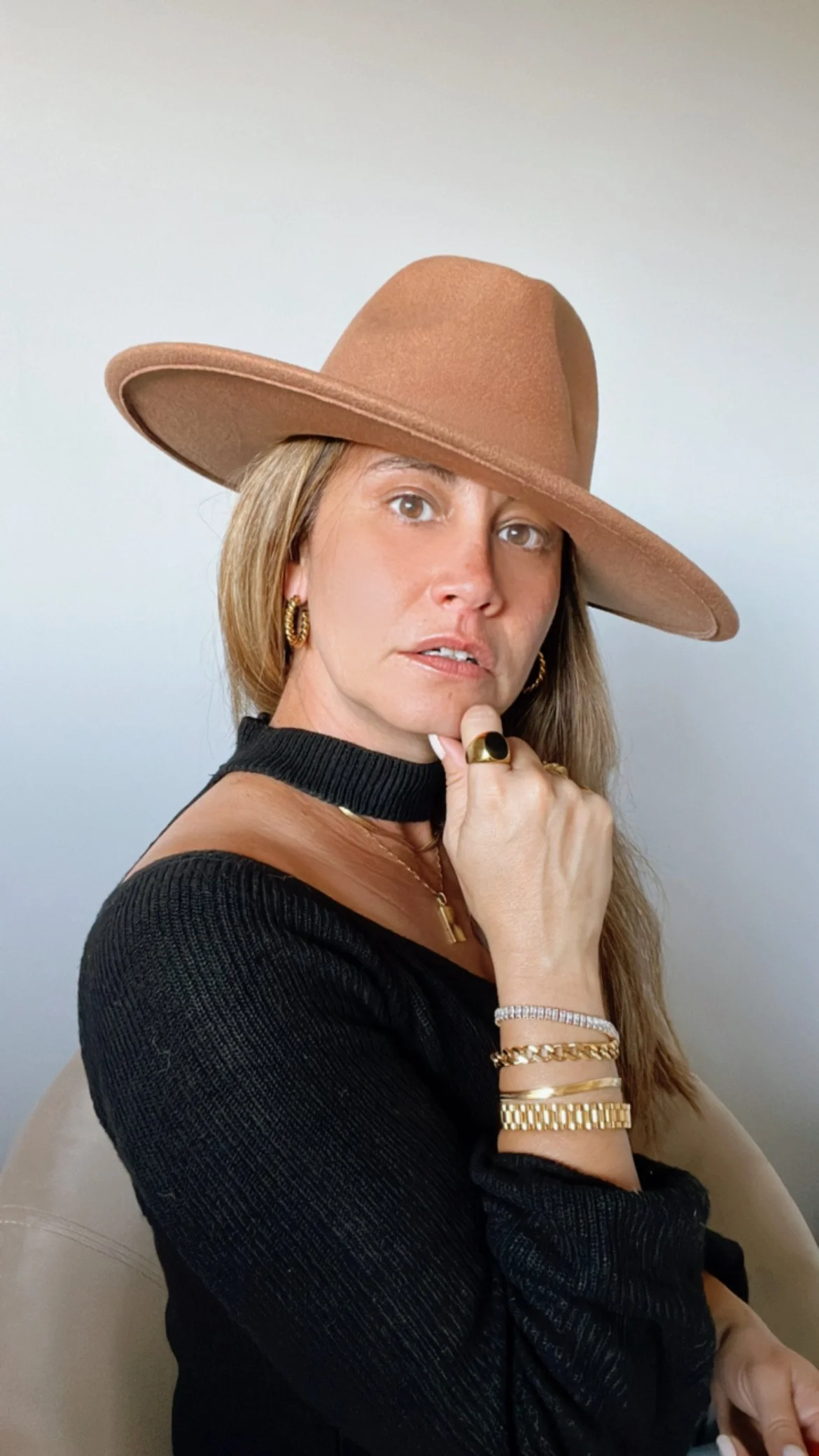 A woman wearing a wide-brimmed brown hat, black sweater, and various gold jewelry, posing with her chin resting on her hand, in front of a plain white background.