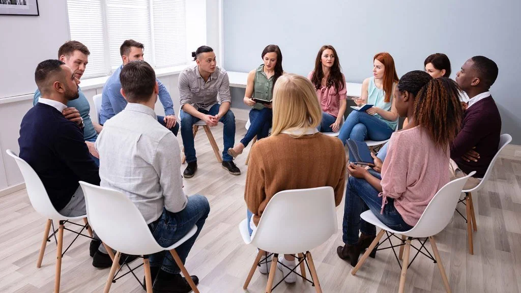 Group of people sitting in a circle during a discussion or meeting in a bright room with large windows and a white brick wall.