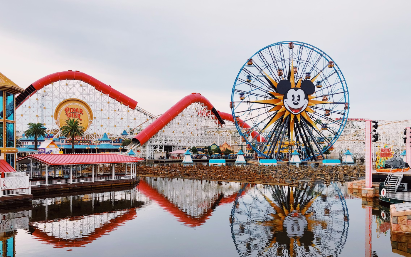 A view of Disney California Adventure Park at Disneyland with a Ferris wheel featuring Mickey Mouse's face, a red roller coaster, and water reflections in the foreground.