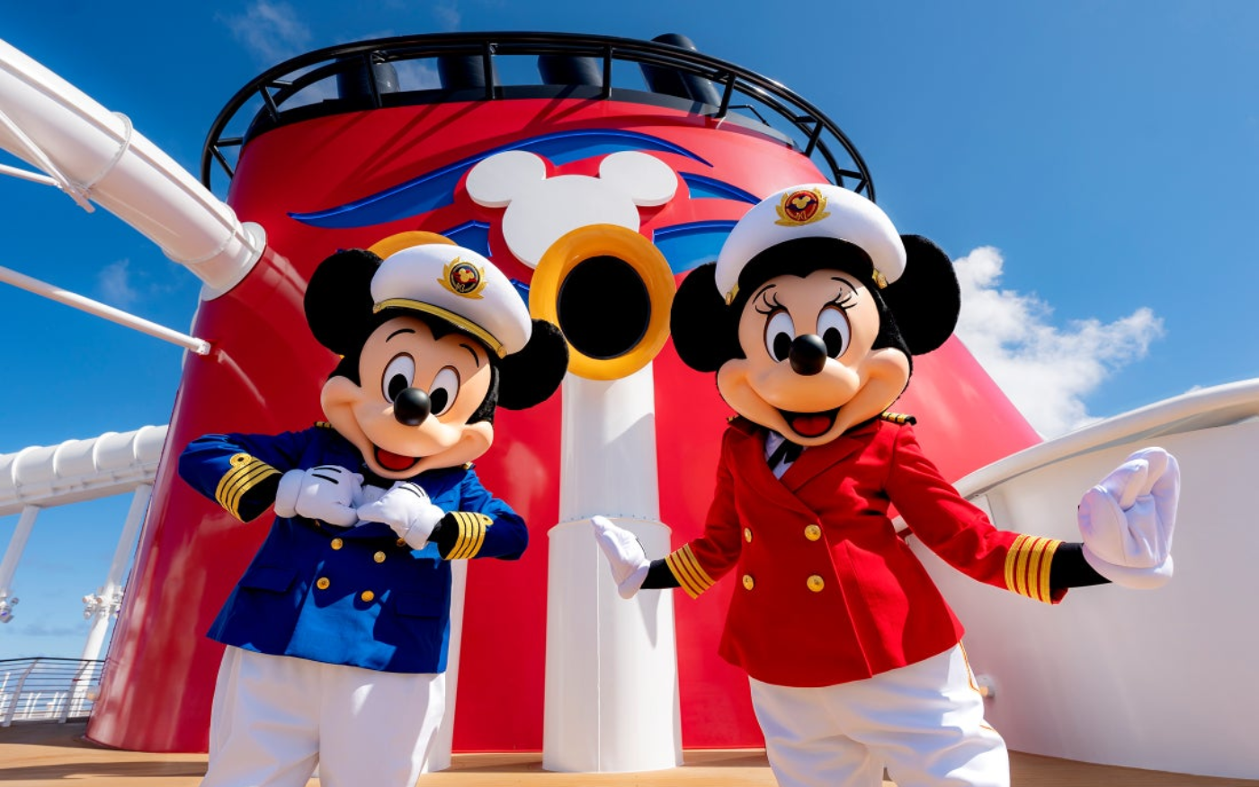 Mickey Mouse and Minnie Mouse dressed as cruise ship officers standing on a cruise ship deck with a large red and white ship funnel, with a blue sky and clouds in the background.