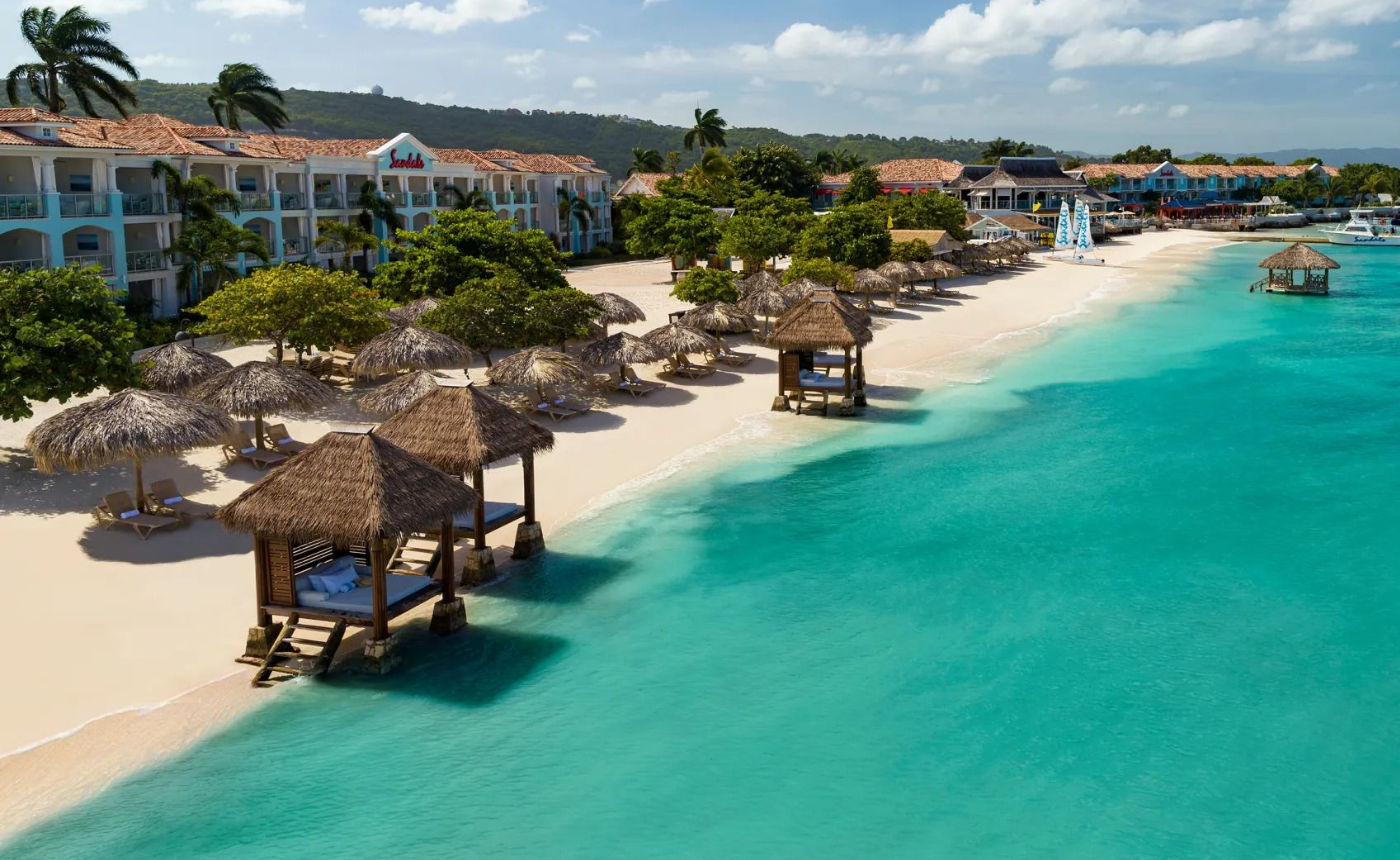 Seaside resort with thatched-roof cabanas, lounge chairs, and hotels along a white sandy beach, turquoise water, and a clear sky.