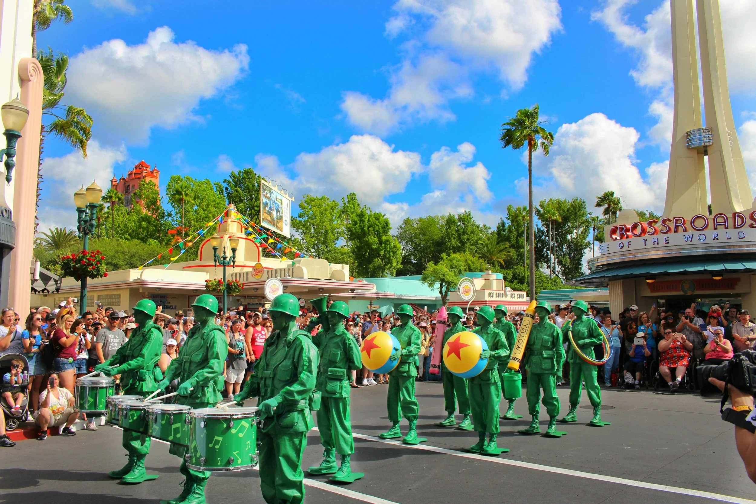 A parade at an amusement park with a marching band dressed in green uniforms, carrying drums, balloons, and a trumpet. Spectators line the street, watching the performance under a partly cloudy sky with palm trees and park buildings in the background.