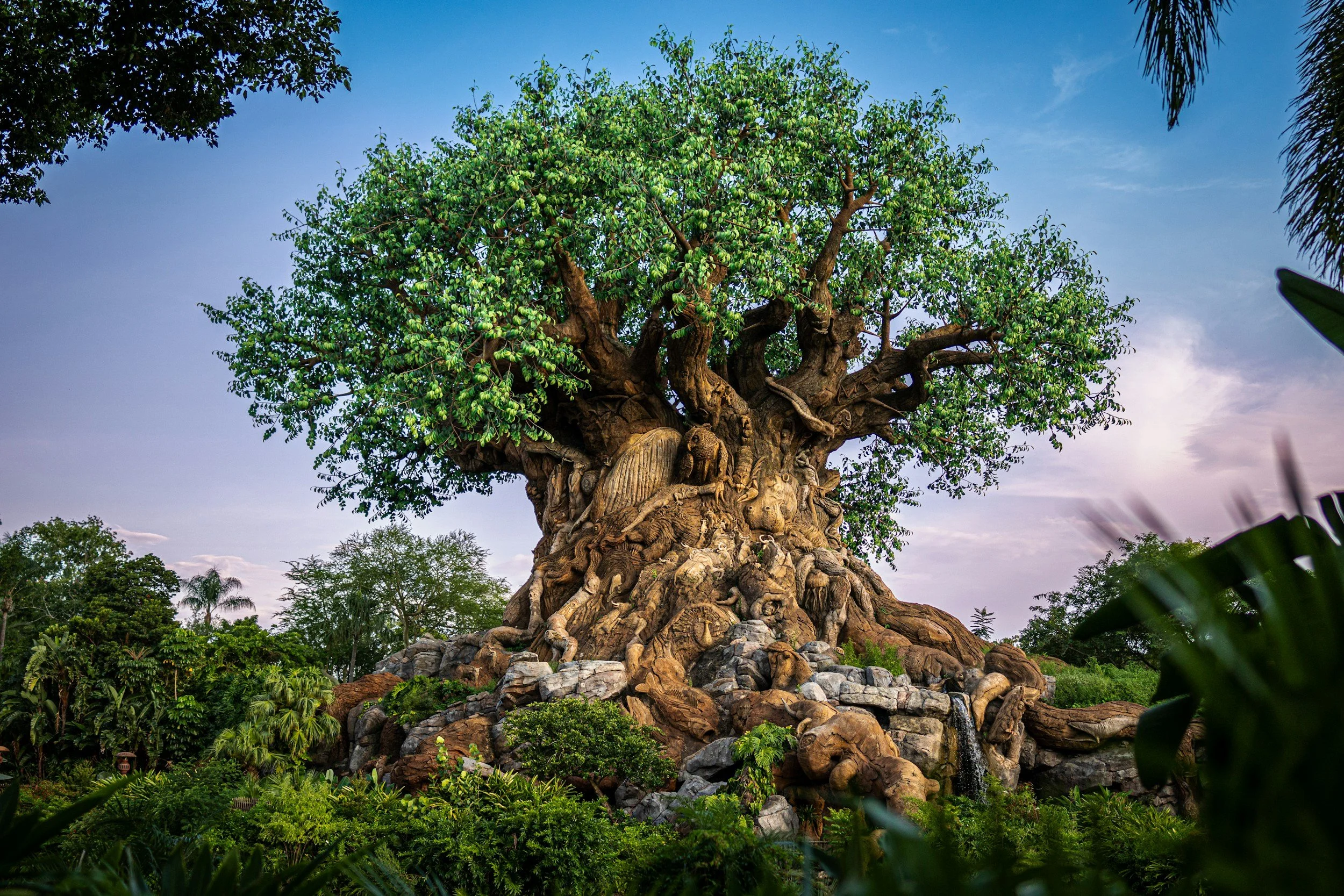 A large artificial tree with carved wooden animals and figures at its base, surrounded by lush greenery and a water feature, under a blue sky.