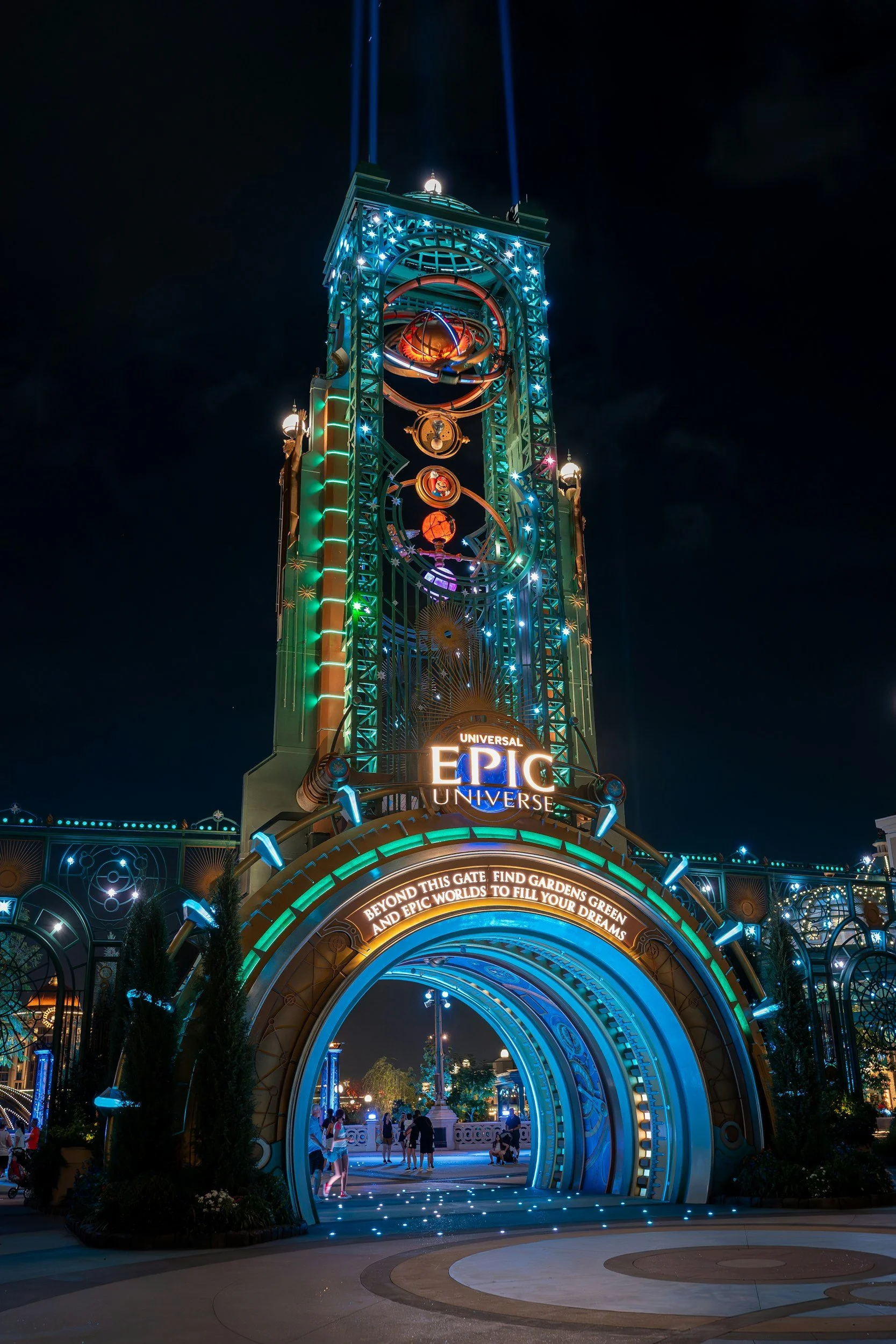 Night view of the entrance to Universal's Epic Universe theme park with a tall, illuminated gate and decorative structure, people walking through the archway.