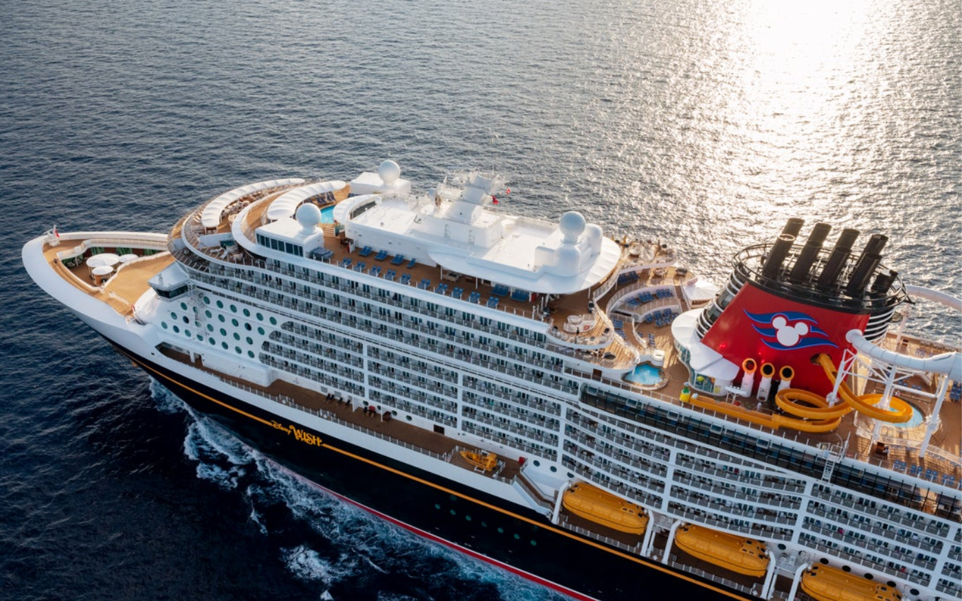 A large cruise ship sailing on the ocean, viewed from above. The ship has multiple decks with pools, lounge chairs, and a water slide at the rear.