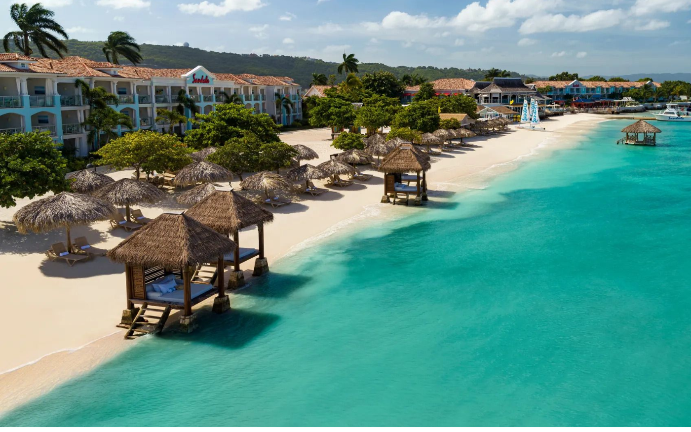 Tropical beach with thatched-roof cabanas, lounge chairs, swaying palm trees, and a resort hotel in the background, adjacent to turquoise ocean water.