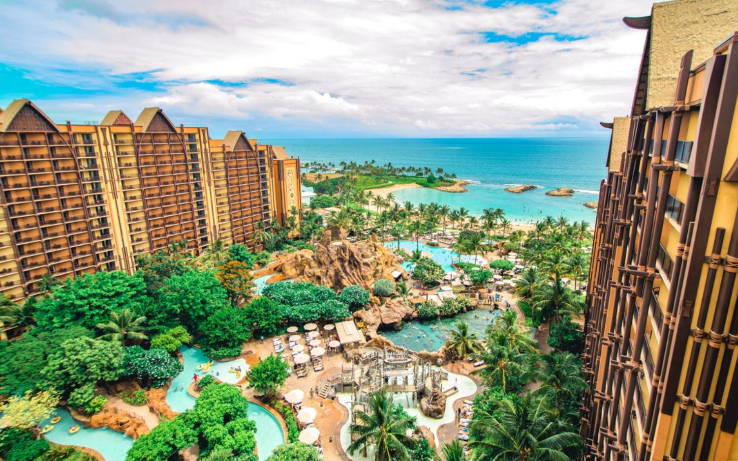 Aerial view of a luxurious beach resort with multiple swimming pools, lush green trees, and tall hotel buildings overlooking the ocean under a partly cloudy sky.
