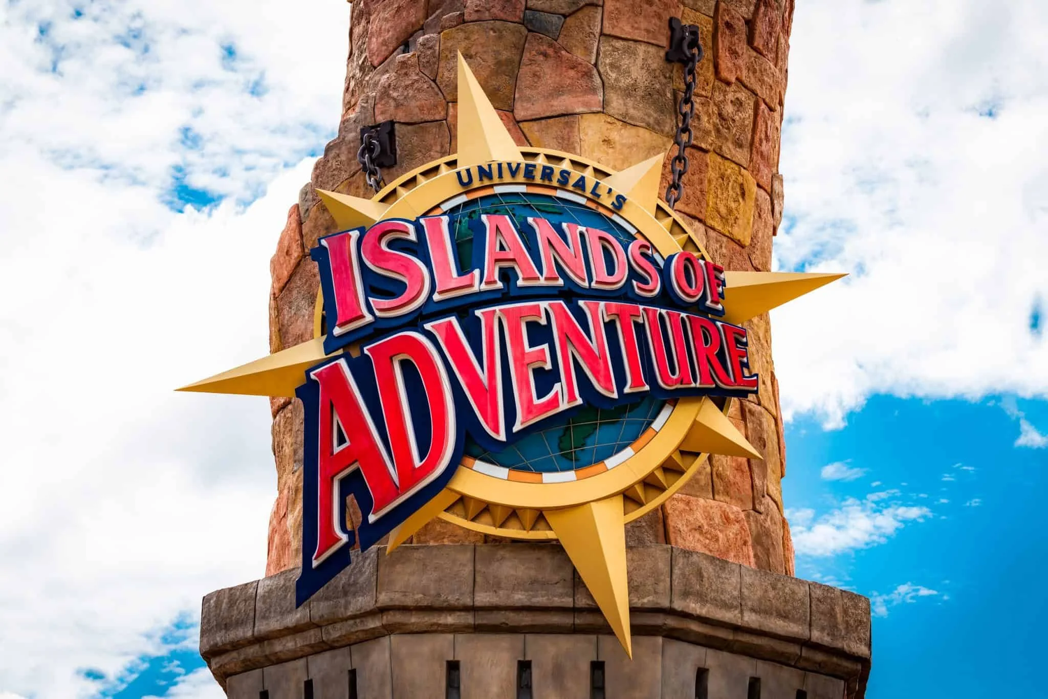 Sign for Universal's Islands of Adventure theme park mounted on a stone tower with a partly cloudy sky background.