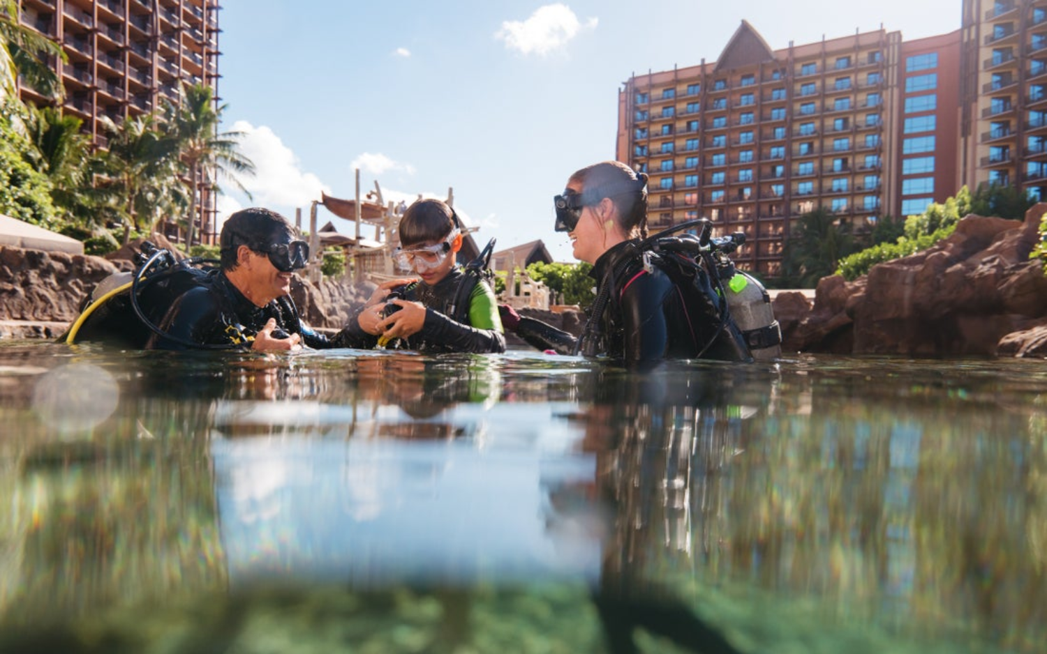 Three people in scuba gear standing in the water, with Aulani Resort in the background, examining something together.